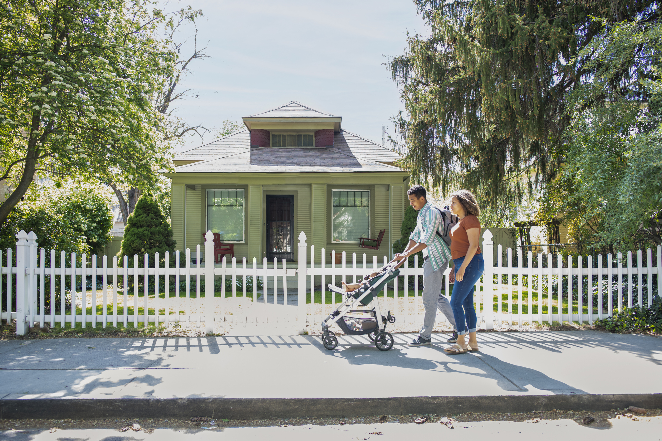 A couple walks on a sidewalk with a stroller in front of a quaint house surrounded by a white picket fence
