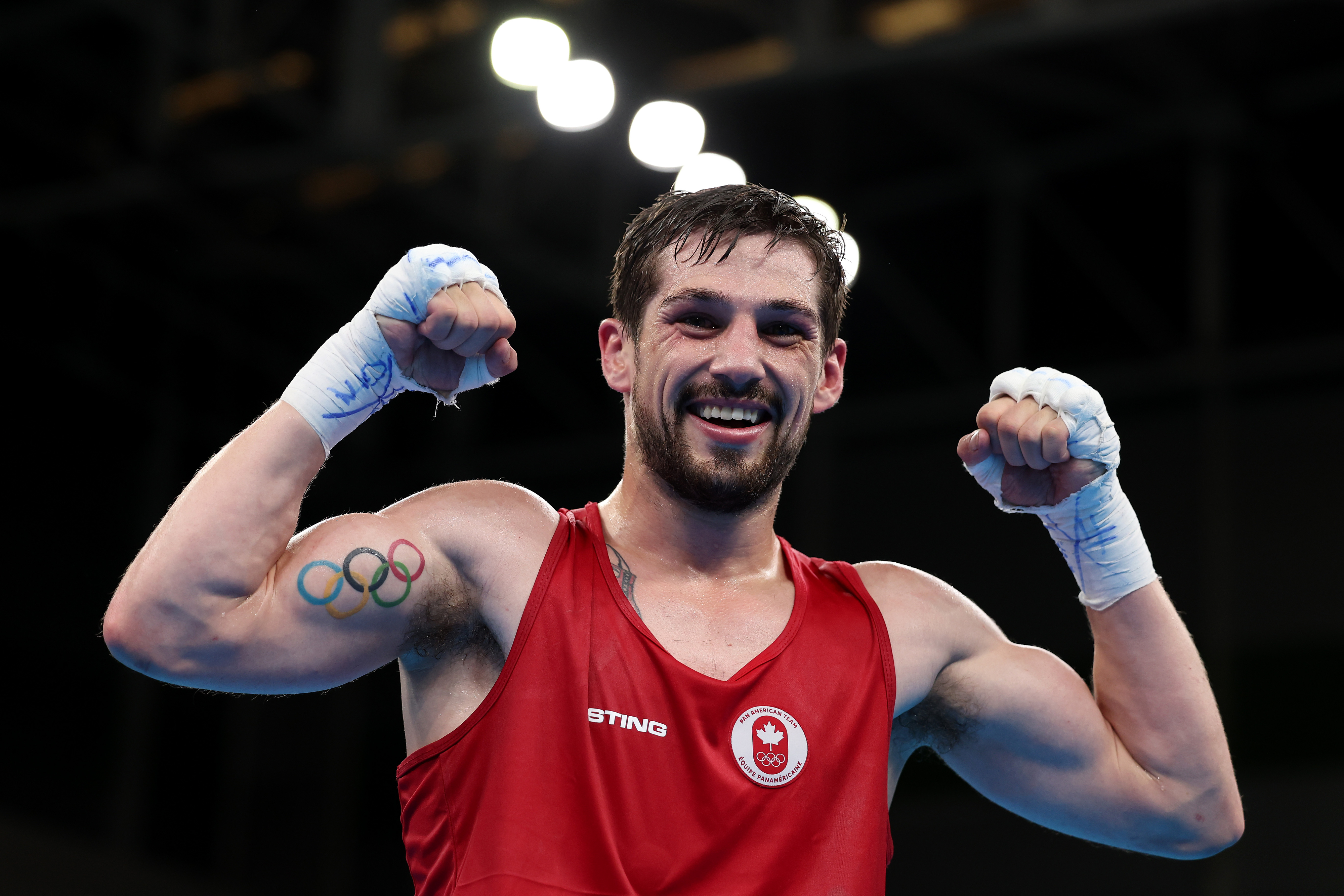 Male boxer in a red tank top celebrates with fists raised. Olympic rings tattoo on left arm