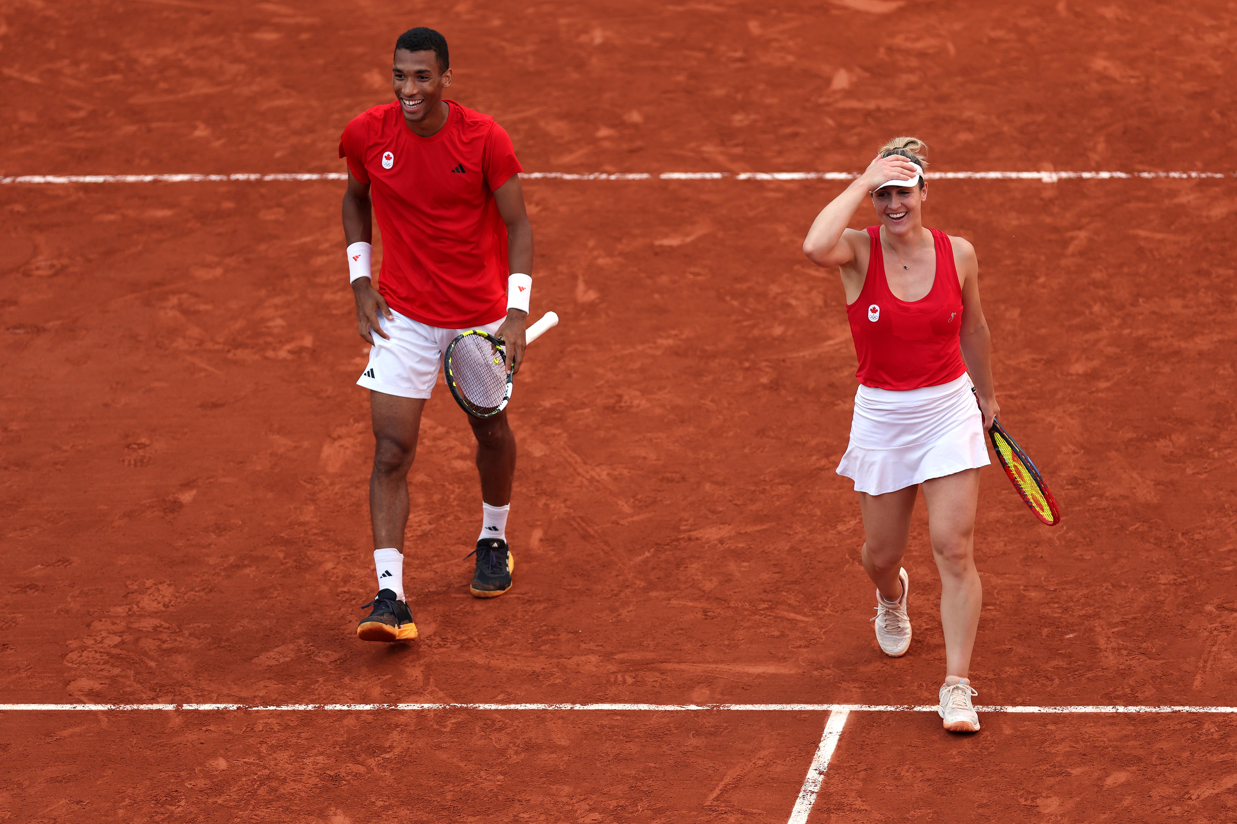 In this image, two tennis players, Félix Auger-Aliassime and Elina Svitolina, smile and walk on a clay tennis court holding their rackets