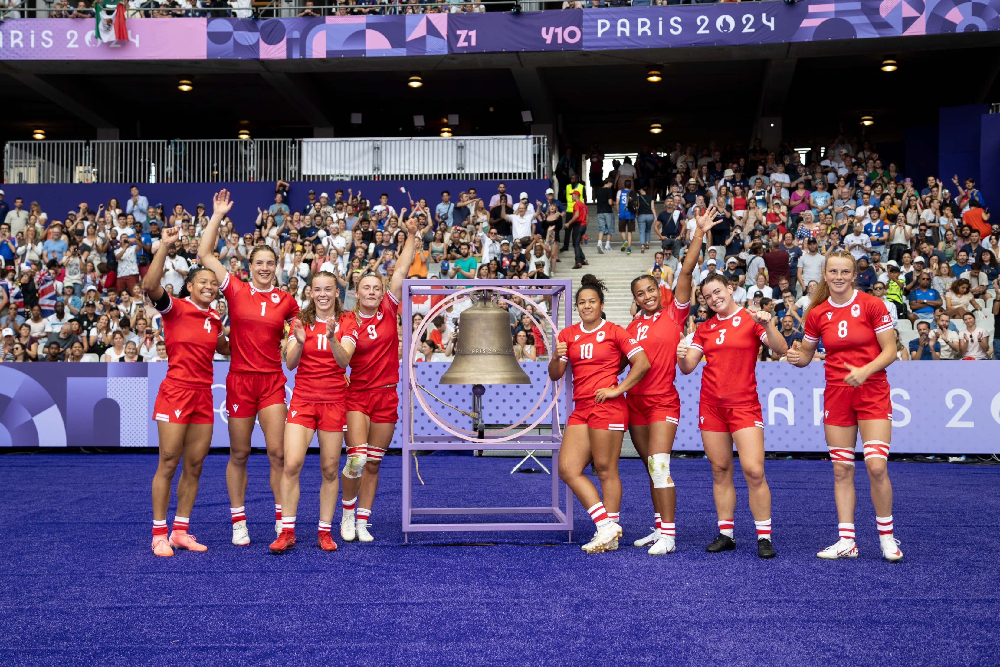 Team of female rugby players, dressed in athletic uniforms, celebrates in front of a crowd and Paris 2024 signage. They gather around a large bell