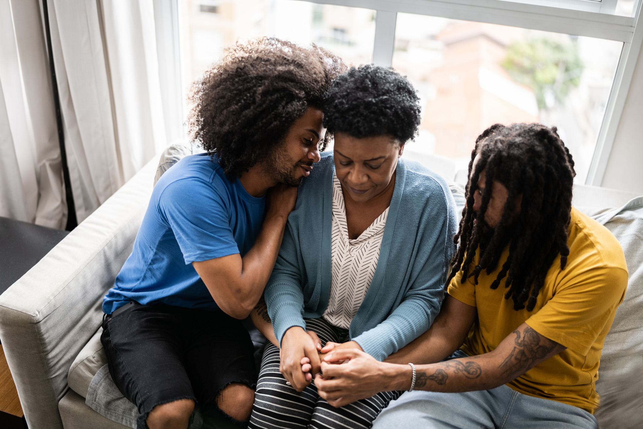 Three people sit closely on a couch, holding hands, appearing to comfort each other