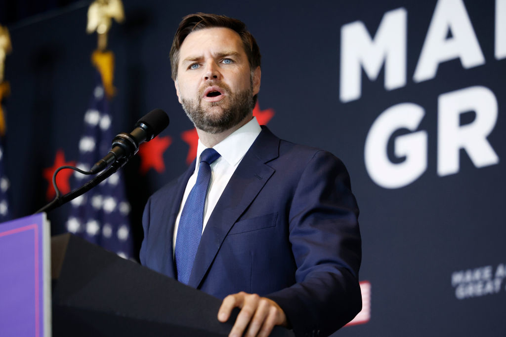 J.D. Vance, dressed in a suit and tie, speaks at a podium with the backdrop displaying the text "MAKE GREAT." Several U.S. flags are visible in the background