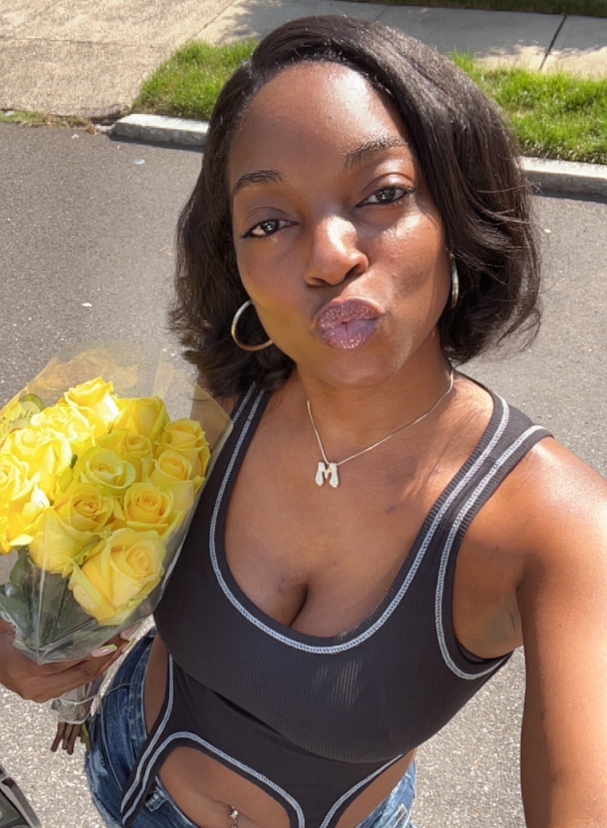 A woman wearing a black tank top and shorts holds a bouquet of yellow roses while taking a selfie outdoors. She is making a kissy face and has shoulder-length hair