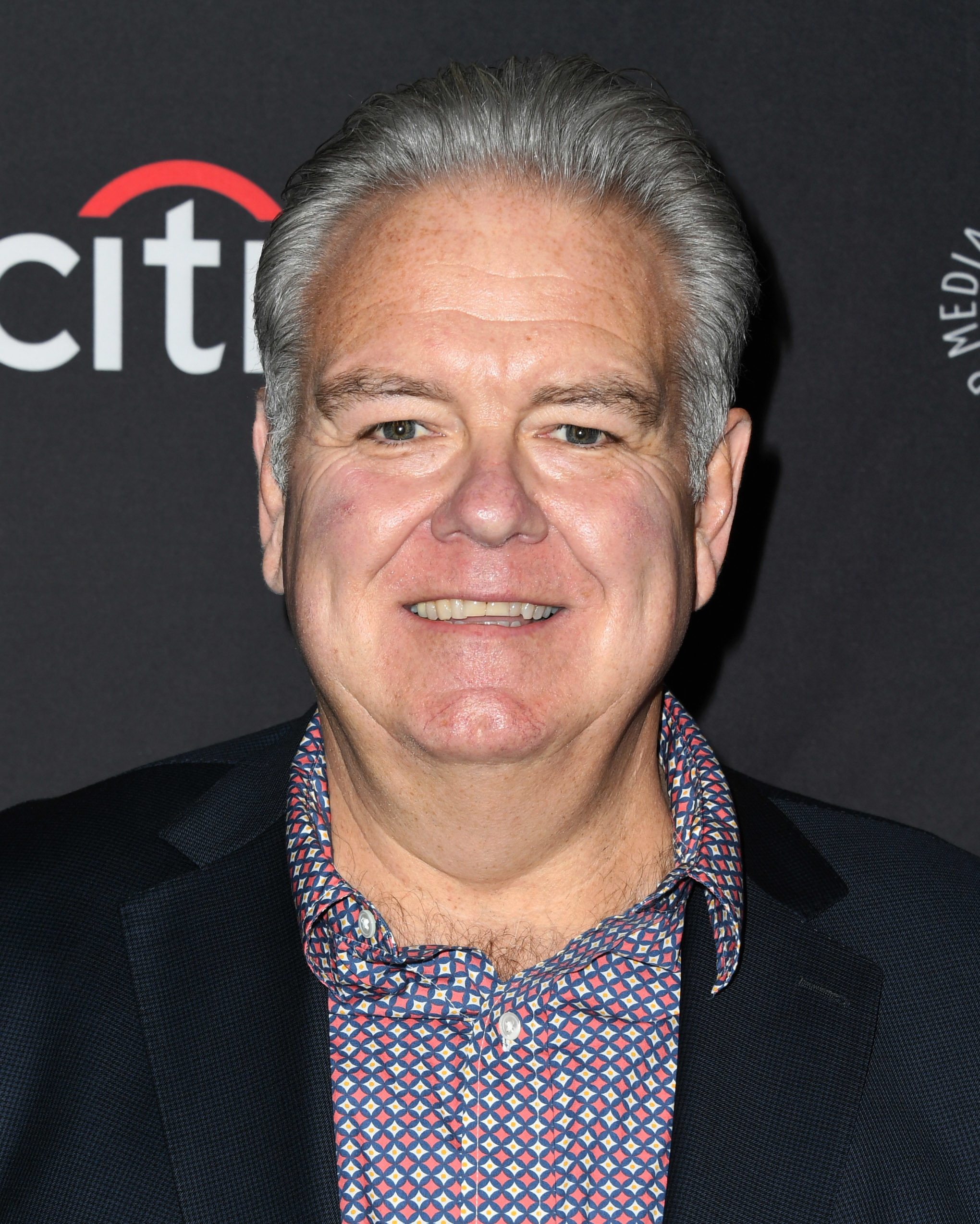 Jim O'Heir smiles at the camera at an event. He wears a patterned shirt and a dark blazer