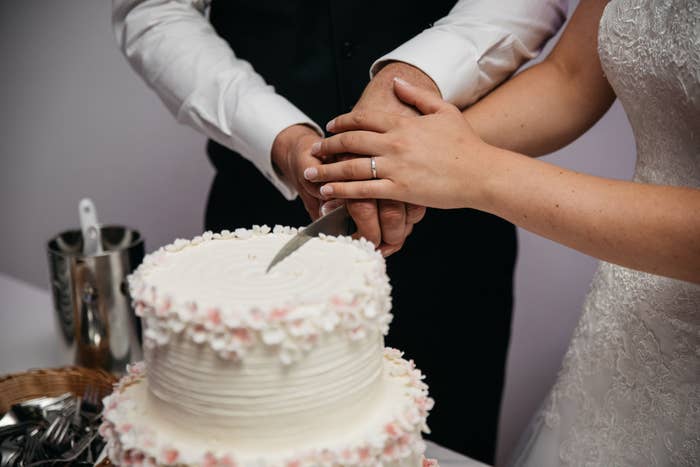 Close-up of a bride and groom cutting a white wedding cake with pink accents, both holding the knife together. No names provided