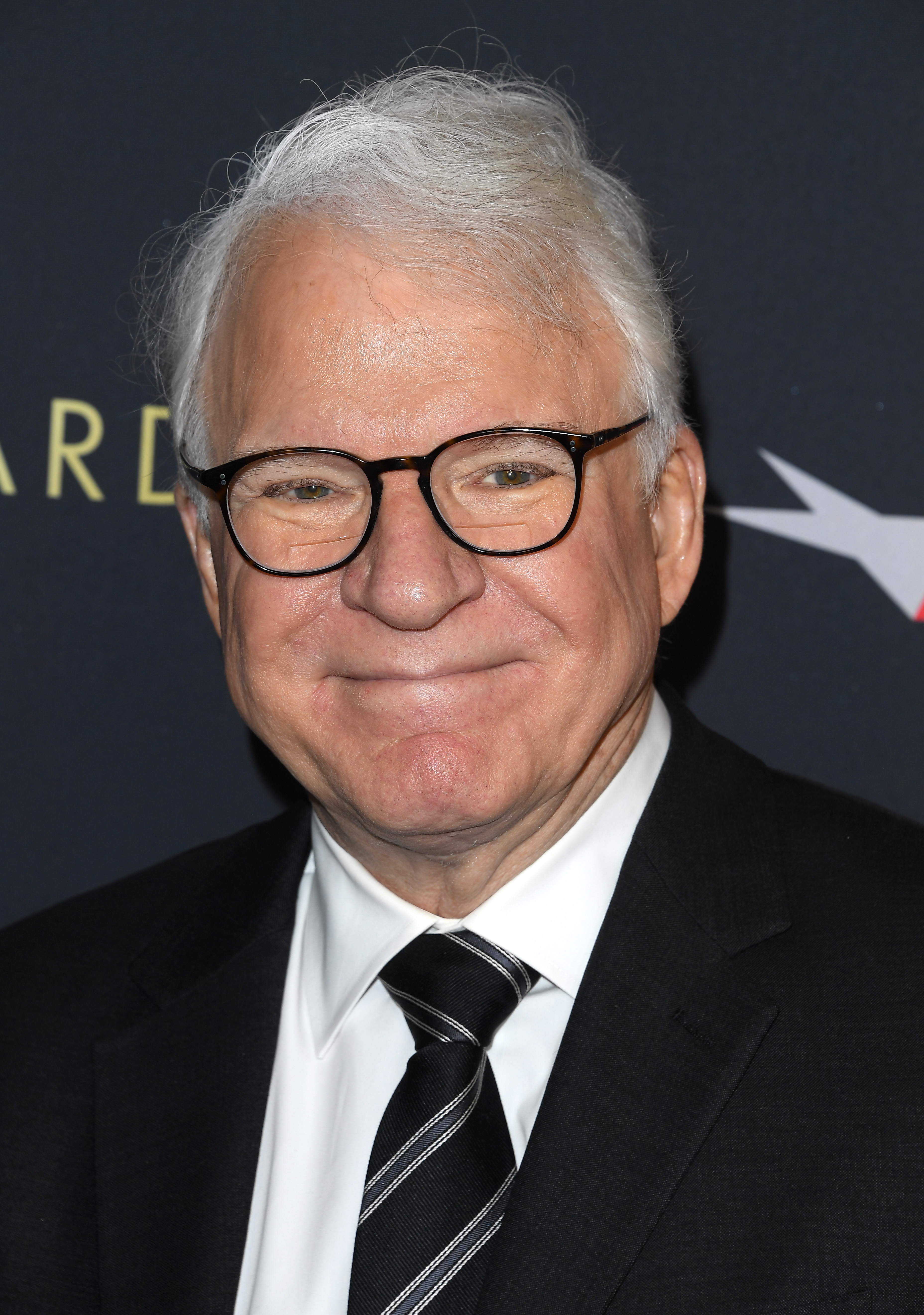 Steve Martin in a black suit and a striped tie, smiling on the red carpet