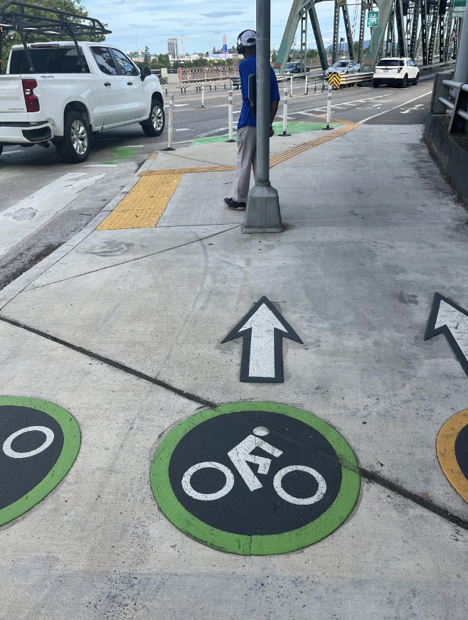 Sidewalk and bike lane with directional signs pointing forward, a person in a blue shirt and helmet waits at the crosswalk near a vehicle and a bridge
