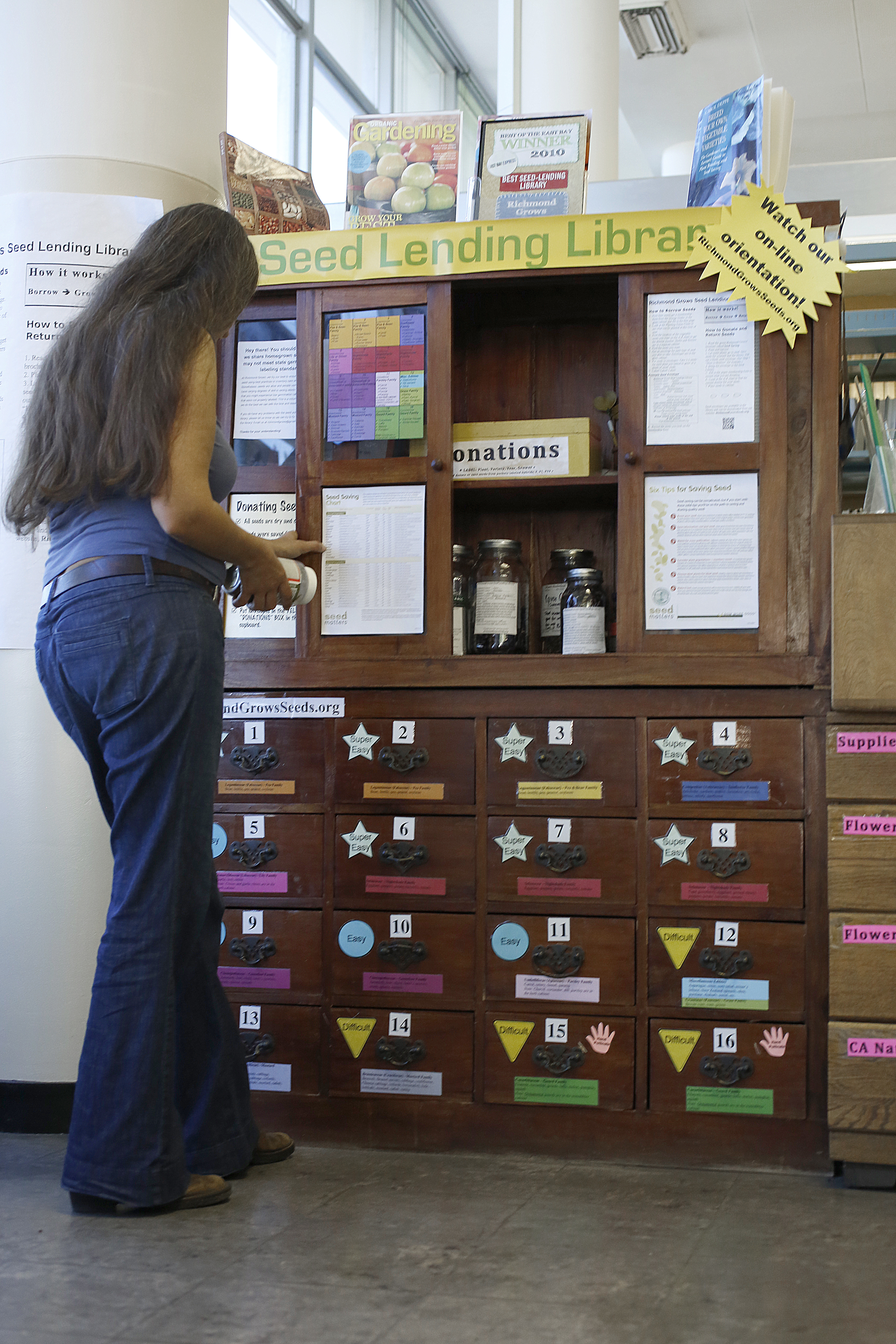A woman looks at a Seed Lending Library cabinet with drawers labeled for different seeds. Signs on the cabinet offer information and instructions