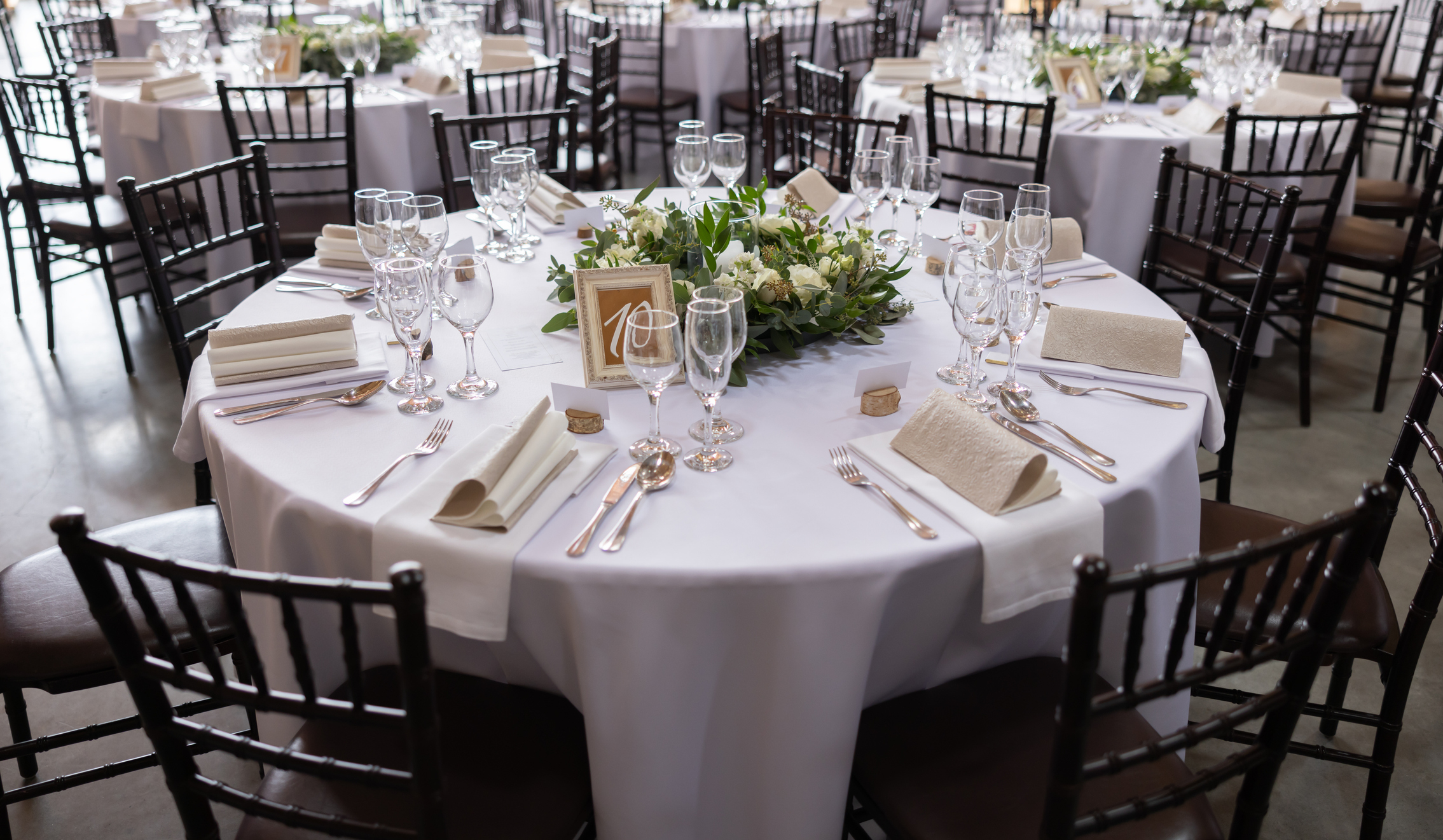 Elegant wedding reception table set with white linens, glassware, cutlery, and floral centerpiece. Multiple tables and chairs surround the setup