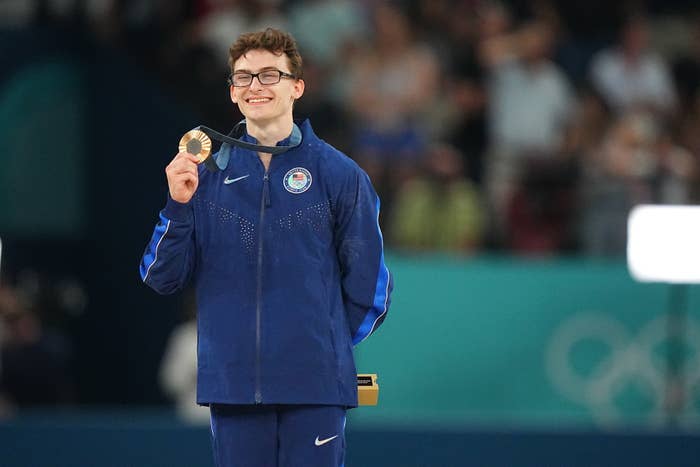 Paralympian Thomas Young holds up his gold medal while smiling during an indoor award ceremony. He is wearing a track suit