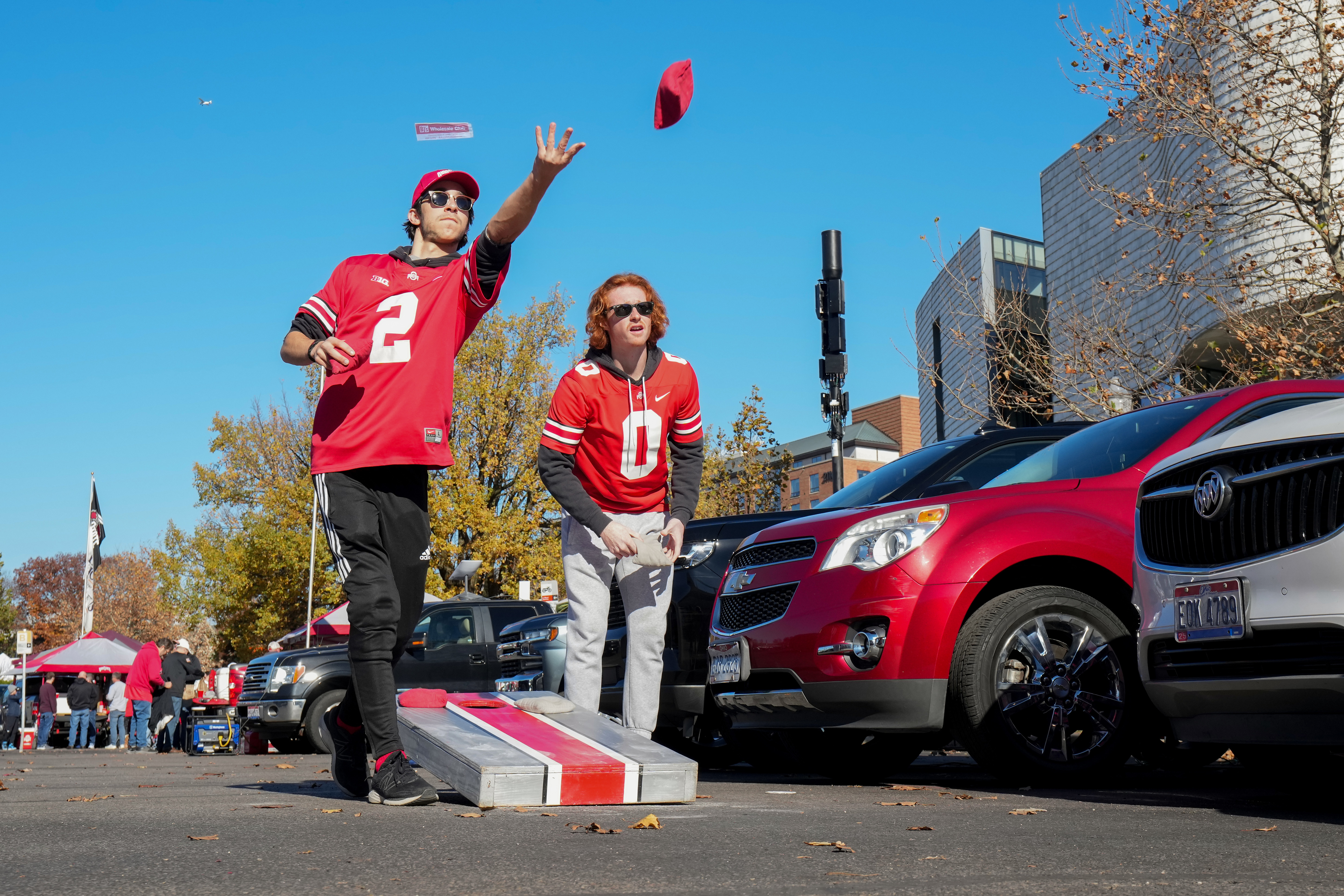 Two men in sports jerseys play cornhole in a parking lot near cars. One man, mid-throw, wears a cap and jersey number 2. The other, jersey number 0