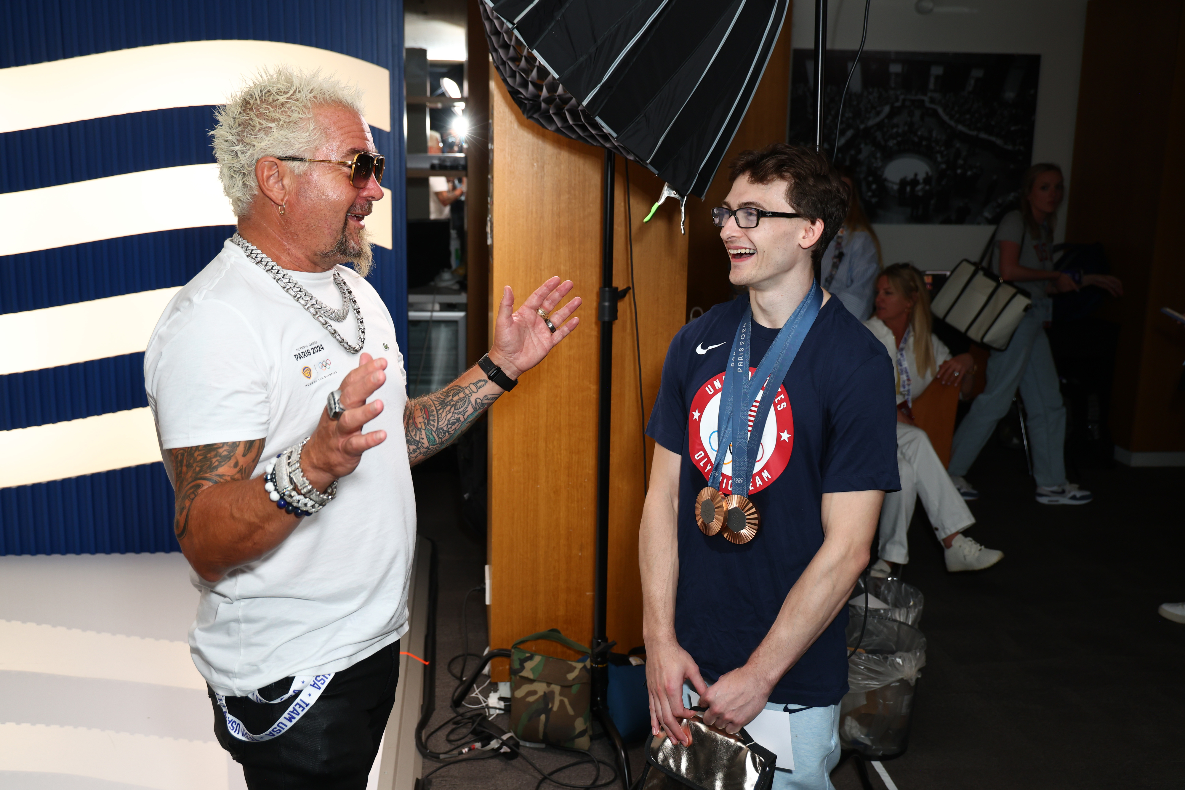 Guy Fieri enthusiastically talks to glasses-wearing man in a navy USA shirt with medals