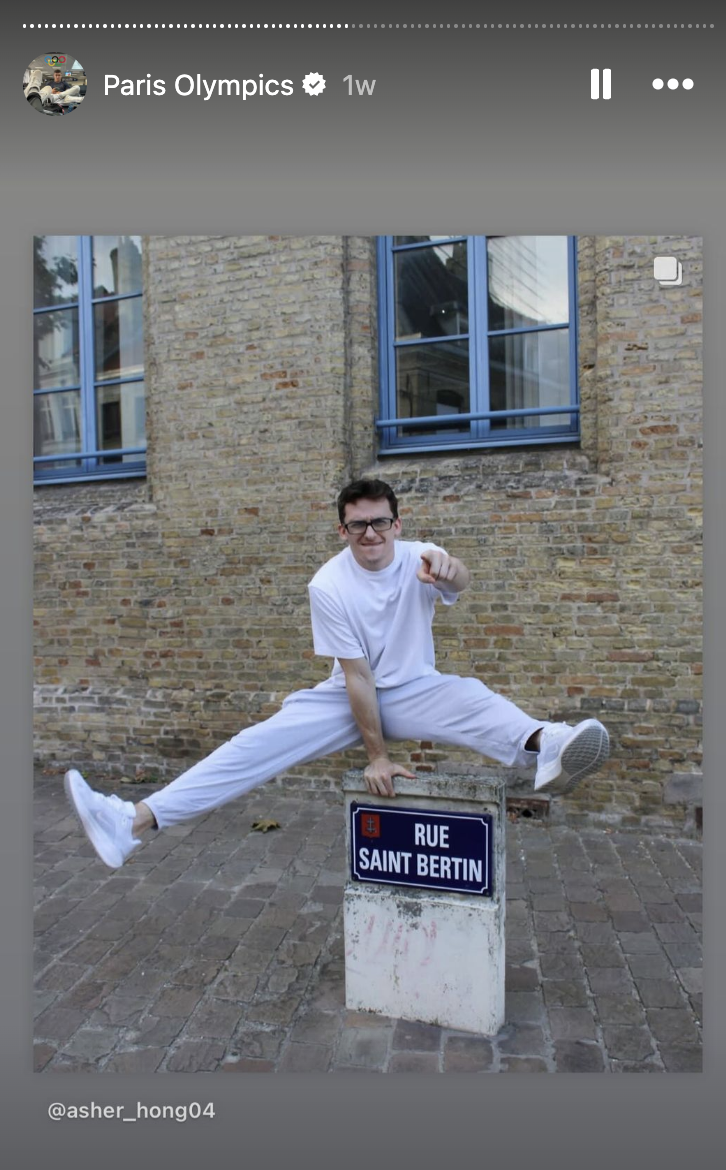 Asher Hong, dressed casually, performs a split jump over a street sign reading "Rue Saint Bertin."