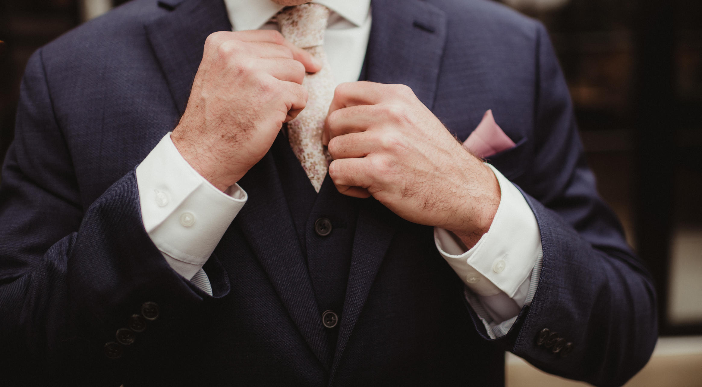 A close-up of an unidentified person adjusting their tie, wearing a formal suit with a vest and a pocket square, typically seen at weddings