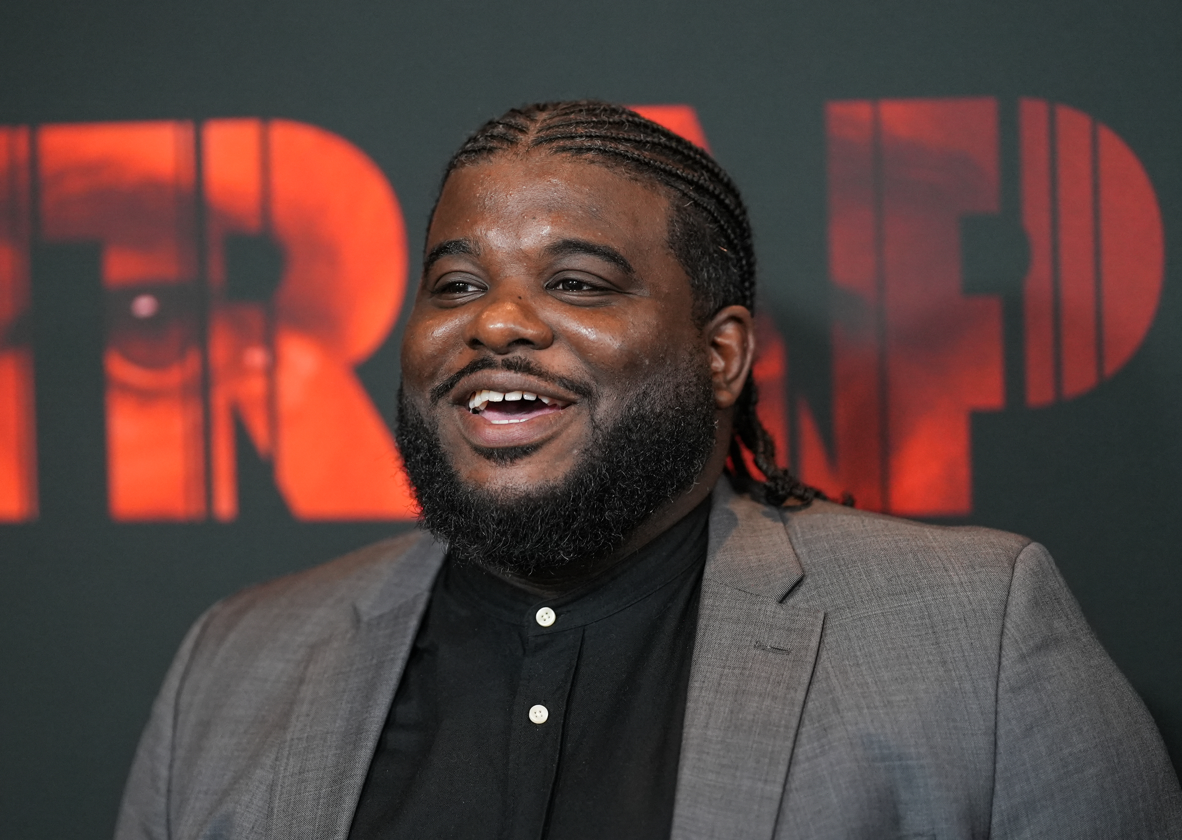 Jonathan Langdon at an event, wearing a gray blazer over a black shirt. Smiling with braided hair, standing against a backdrop with large red letters