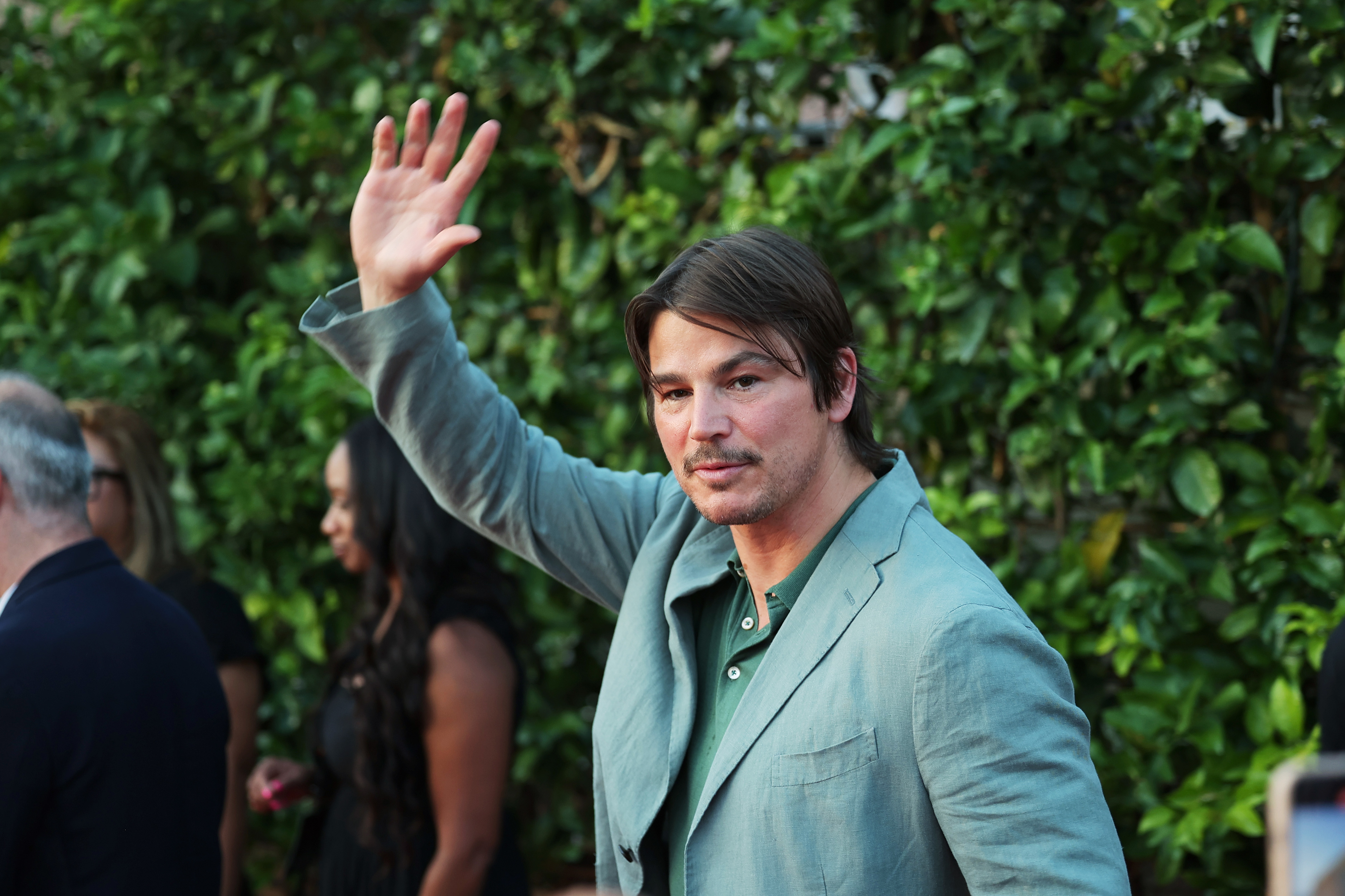 Josh Hartnett, waves at an event outdoors with lush greenery in the background. Other attendees are visible but not clearly identifiable