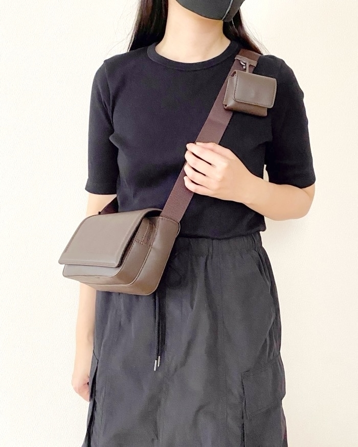 A woman wearing a black face mask, a simple black t-shirt, and a black skirt, carries a brown crossbody bag and a matching small pouch