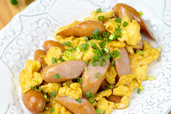 白い皿に盛られたスクランブルエッグとソーセージの料理。緑のネギがトッピングされています。