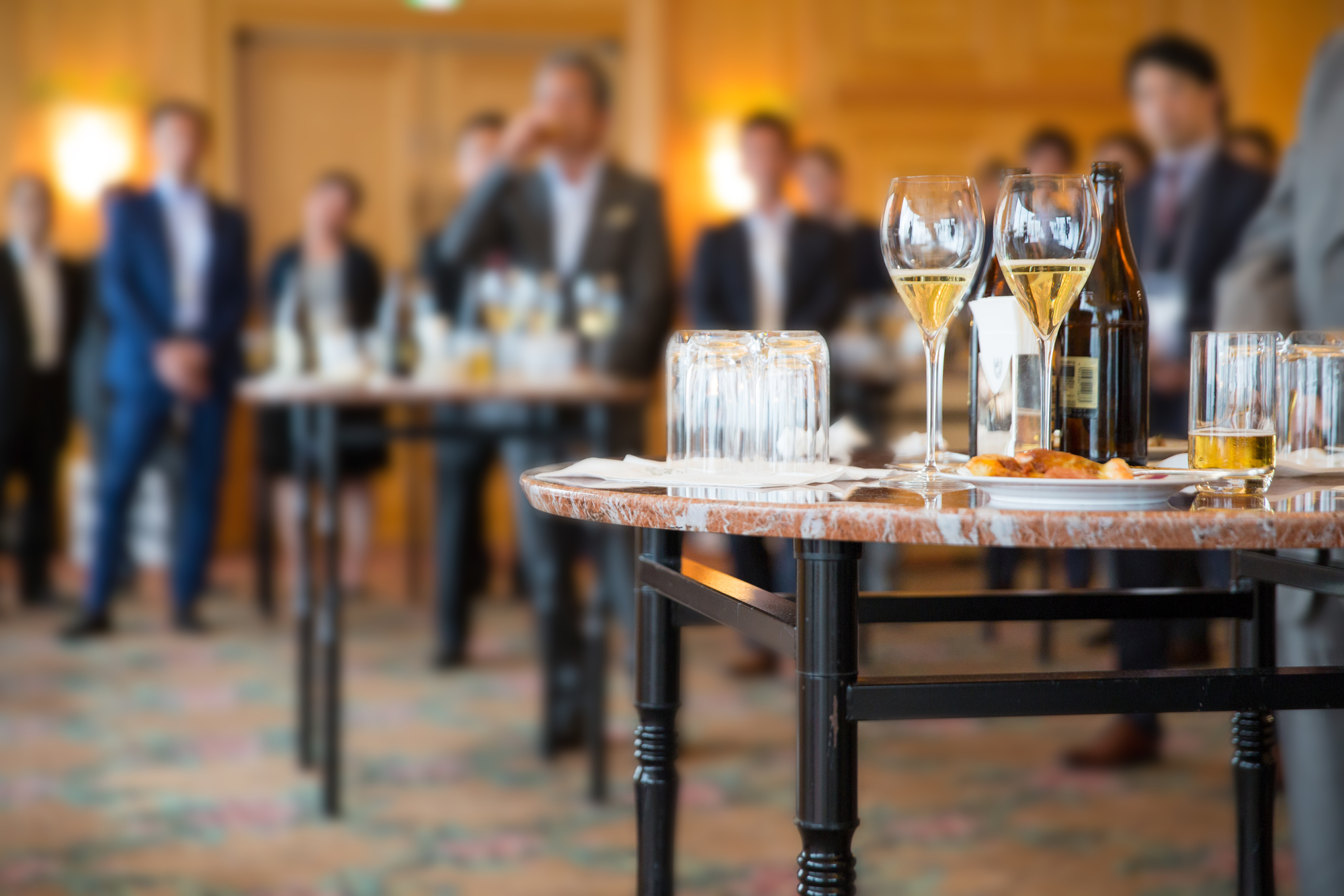 グラスやボトルの並ぶテーブルがあり、背景にフォーマルな服装の人々がぼやけて見えるレセプションの様子。
