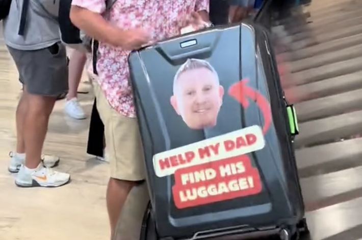 男性が空港の手荷物受取エリアで自分のスーツケースを持ち、バッグには「HELP MY DAD FIND HIS LUGGAGE!」と大きな文字が書かれています。
