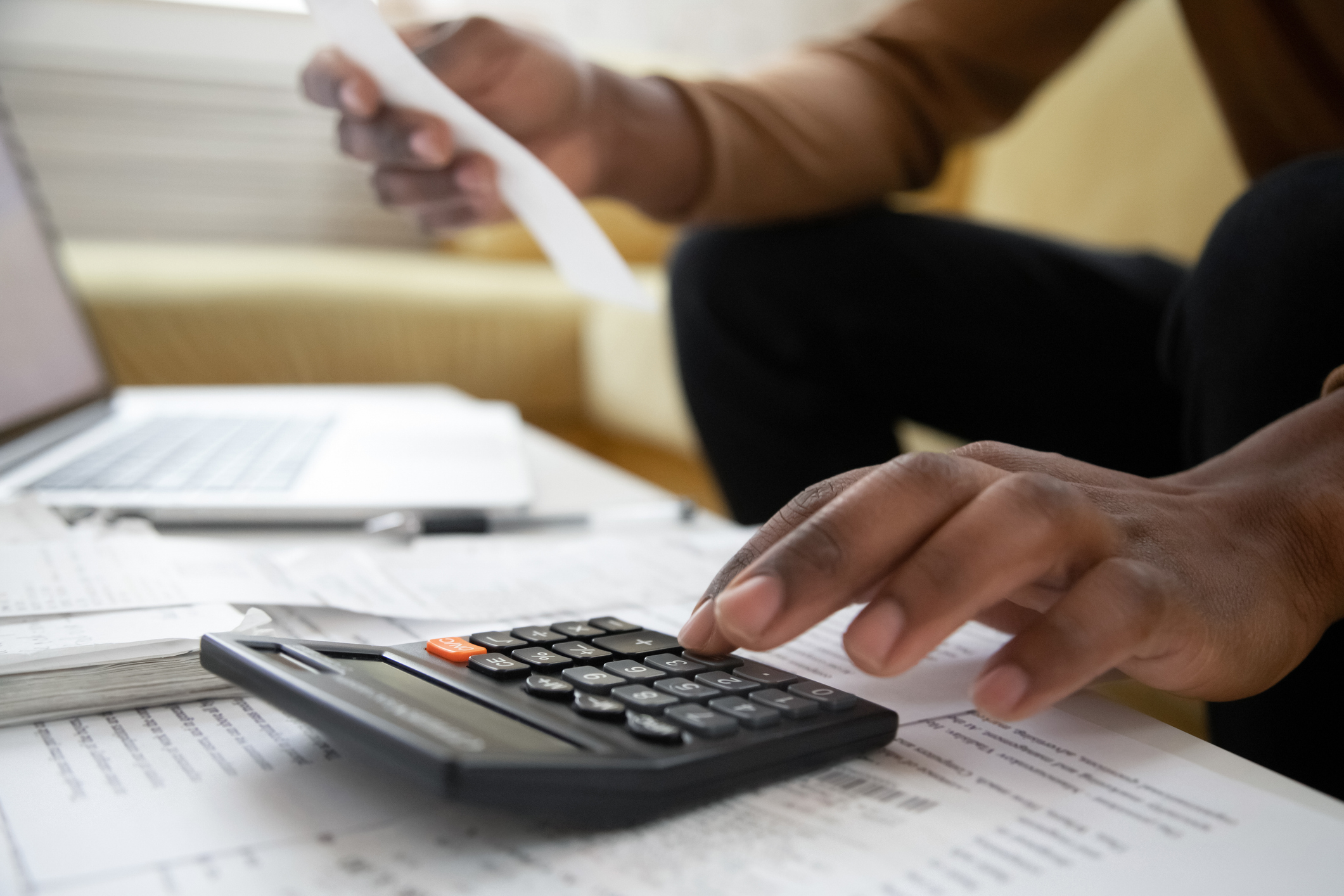 Person using a calculator and holding a receipt, with documents and a laptop on the table