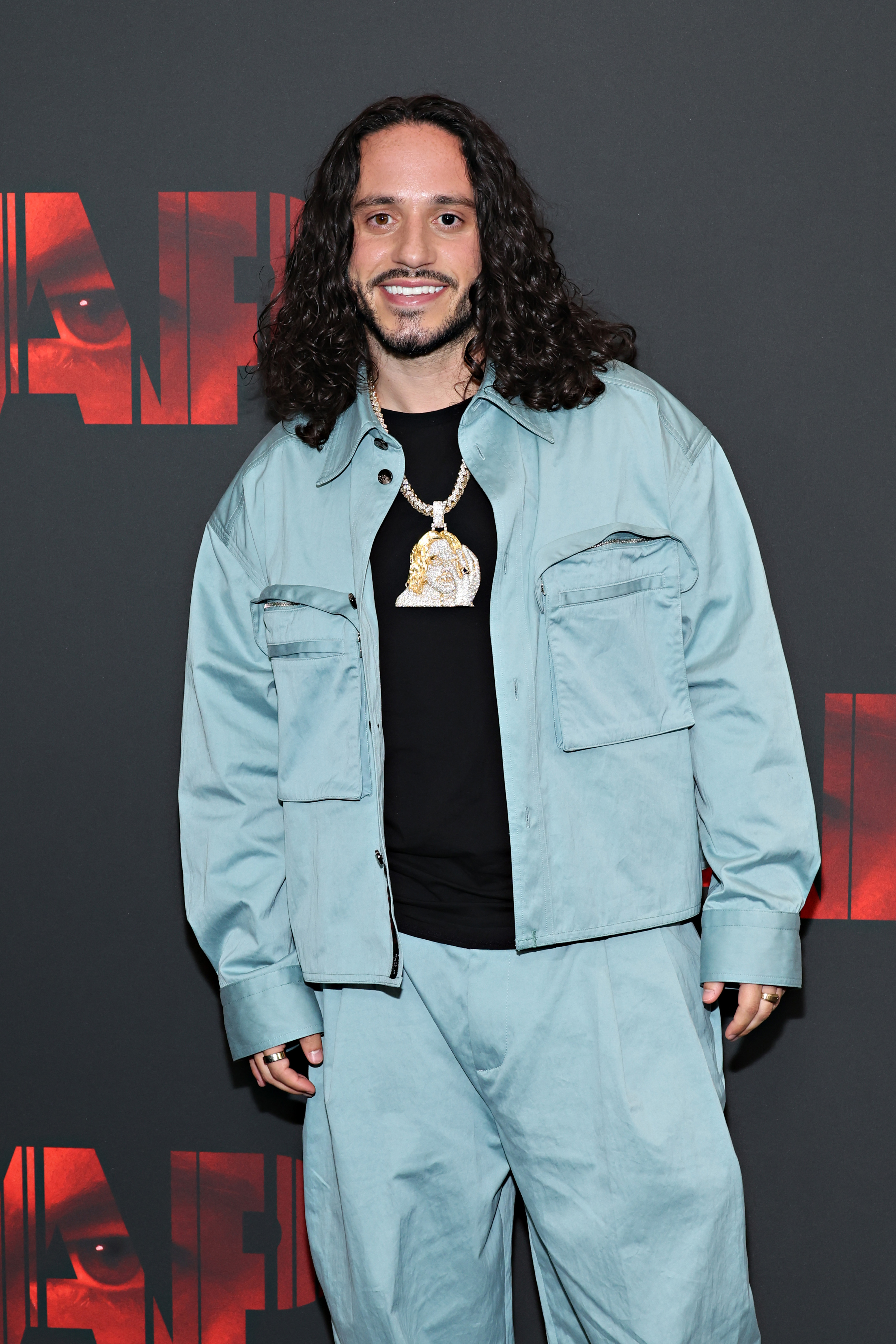 Russ smiles at a red carpet event, wearing a teal jacket and baggy pants, with a large pendant necklace over a black shirt