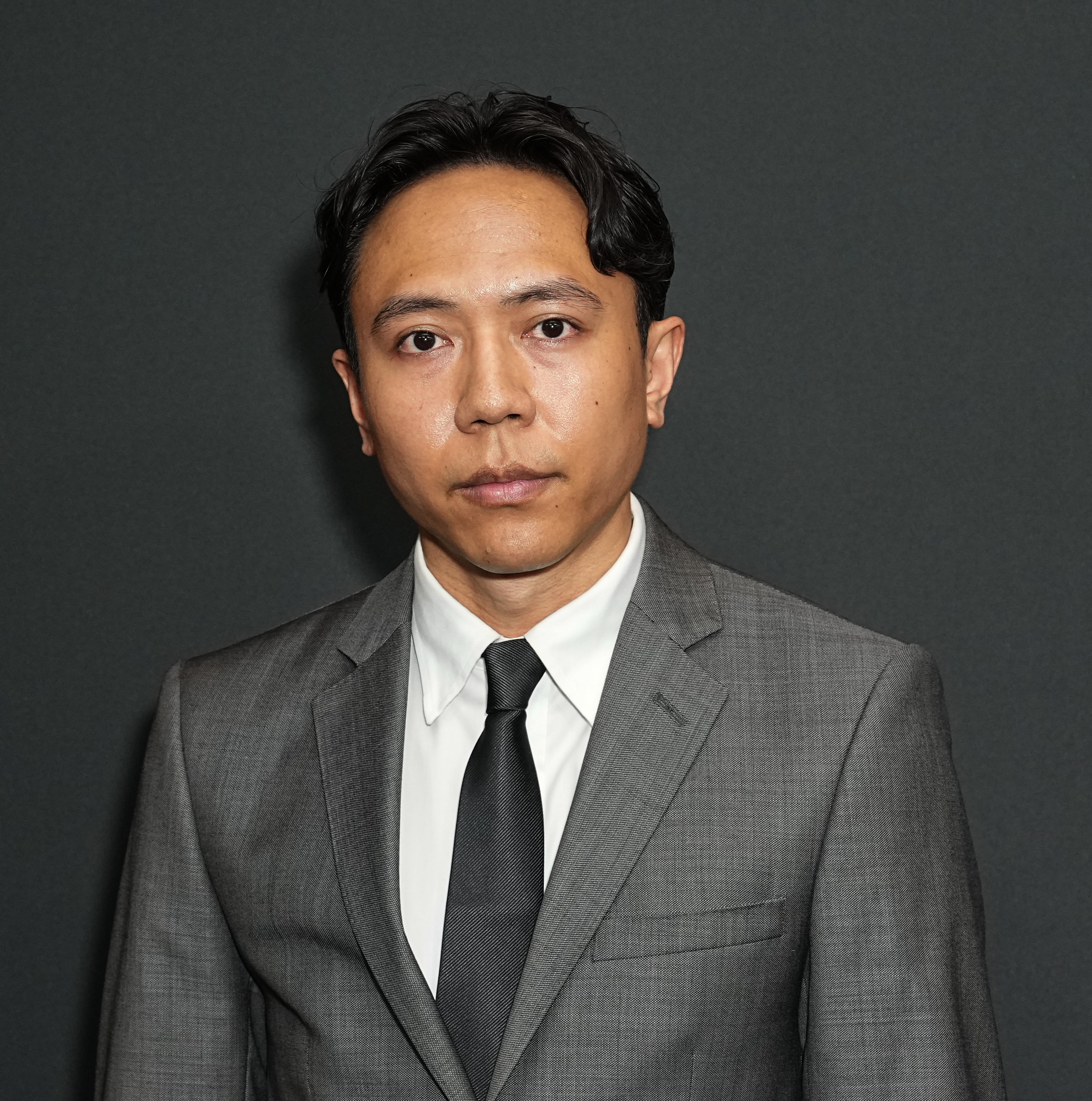Mark Bacolcol in a gray suit, white shirt, and black tie stands against a plain background. His expression is serious