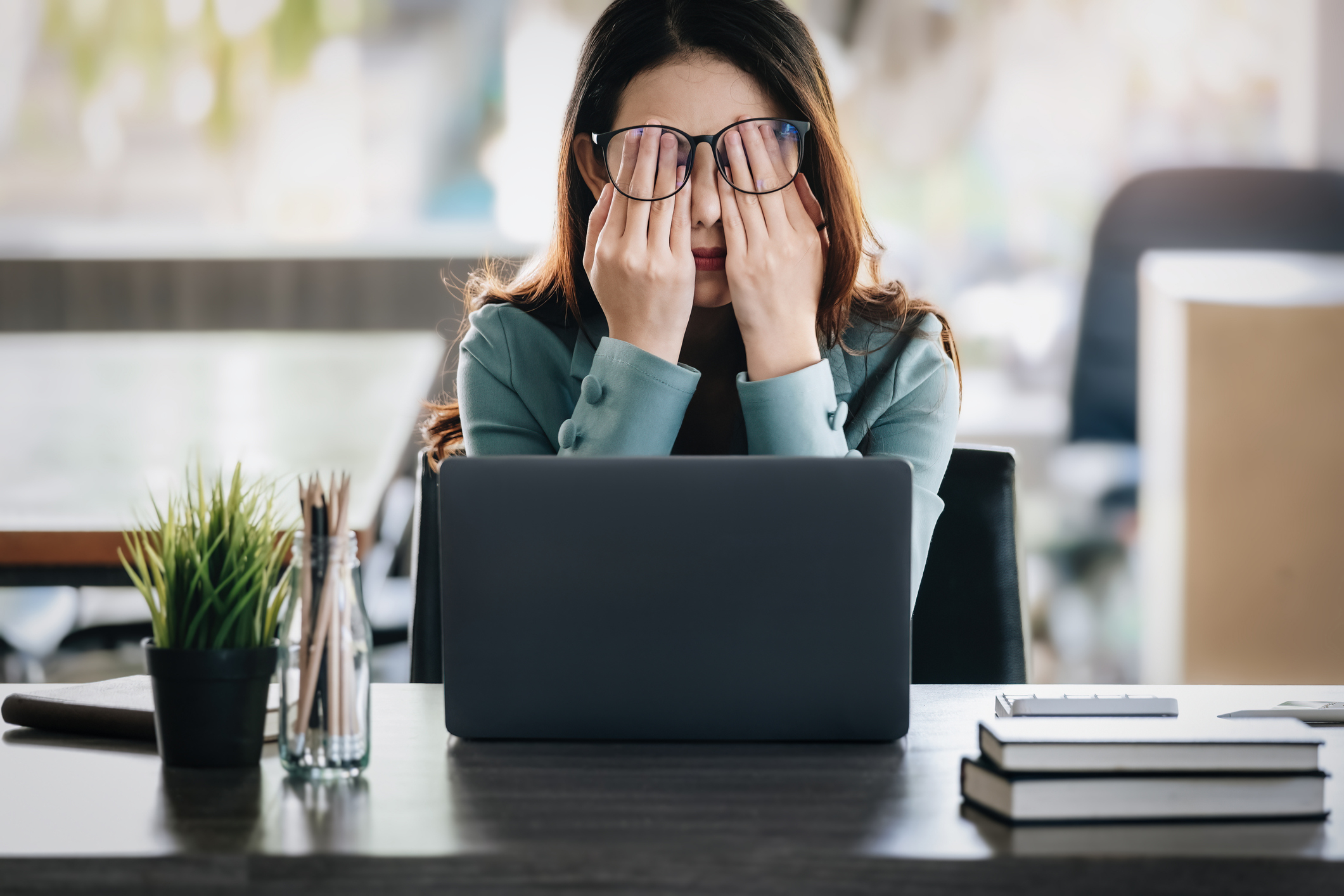 A woman at a desk, sitting in front of a laptop, rubbing her eyes with both hands, looking tired. The desk has a plant, a pen holder, and stacked books