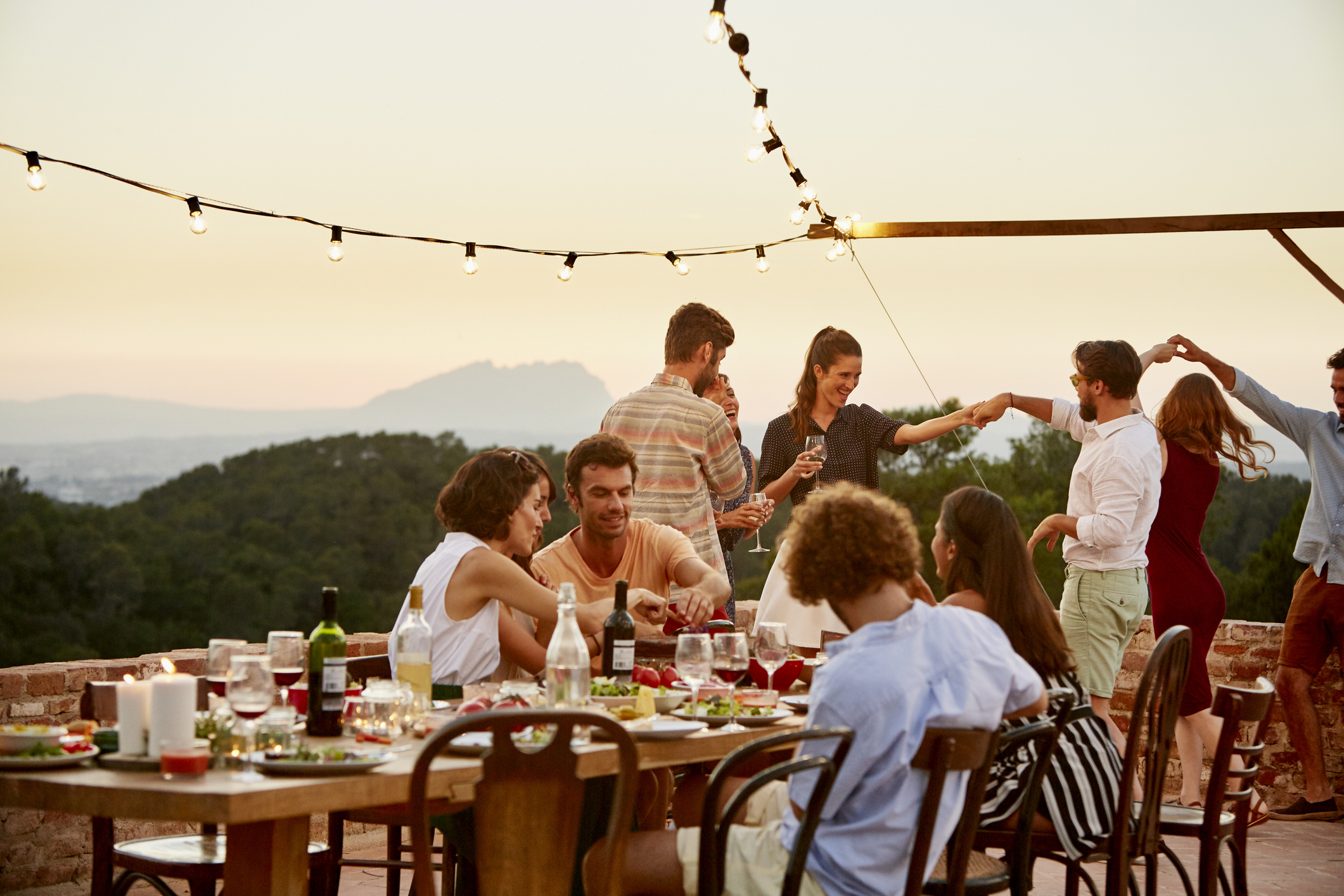 People dining and socializing outdoors at a sunset-lit gathering. The table is set with food, glasses, and wine. String lights are hung above the group