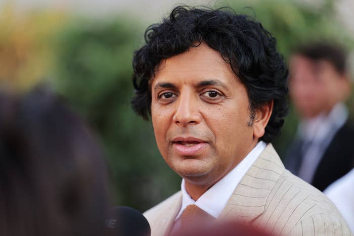 M. Night Shyamalan in a light-colored suit poses for a photo at an outdoor event