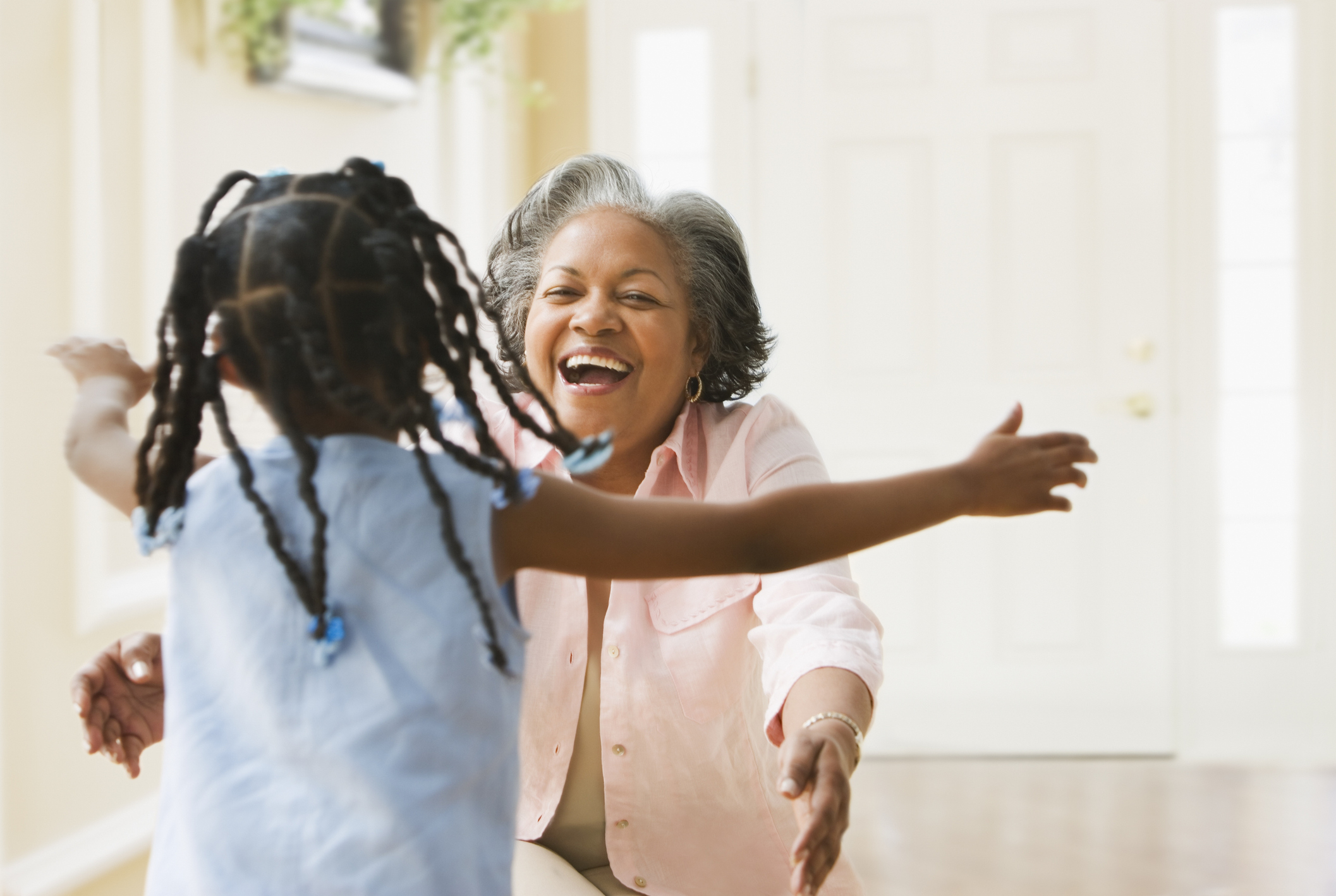 A joyful older woman with short gray hair, sitting and opening her arms to embrace a young girl with braided hair running towards her