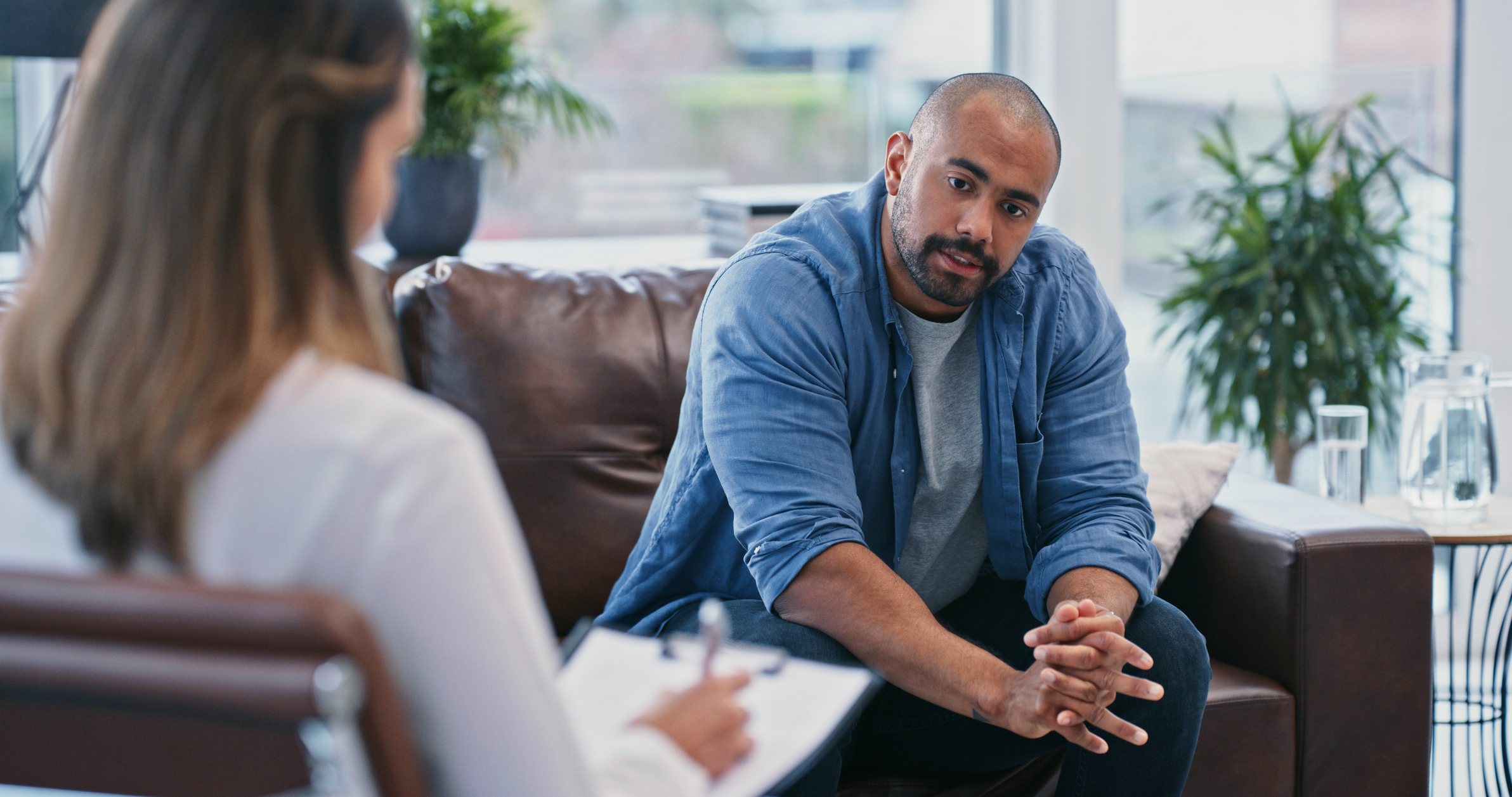 A man in a casual shirt and jeans sits on a couch, talking to a woman taking notes on a clipboard in a modern office setting