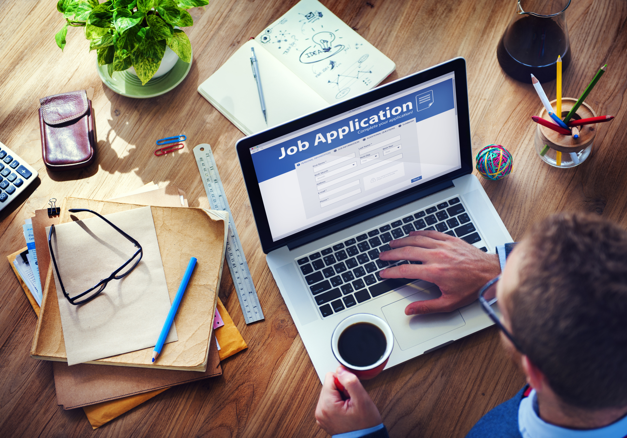Person using a laptop with a job application form on the screen, holding a cup of coffee. Desk covered with papers, ruler, notebook, plant, and other office supplies