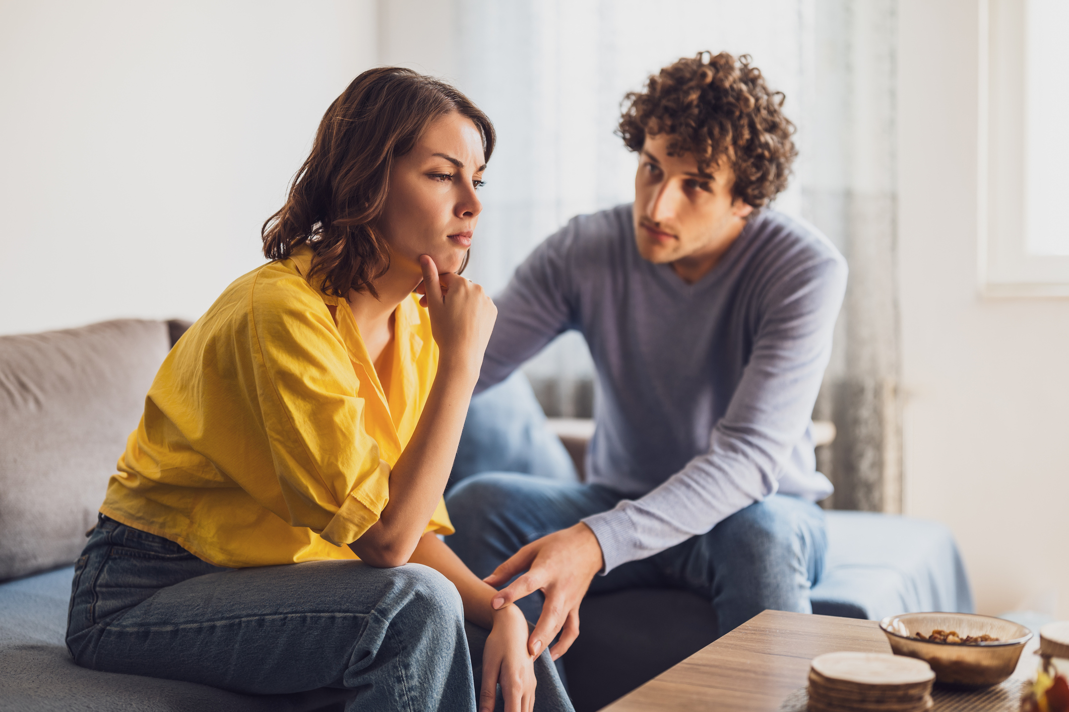 A woman in a casual yellow top sits thoughtfully, while a man in a casual sweater sits beside her, appearing concerned. They are on a couch in a home setting