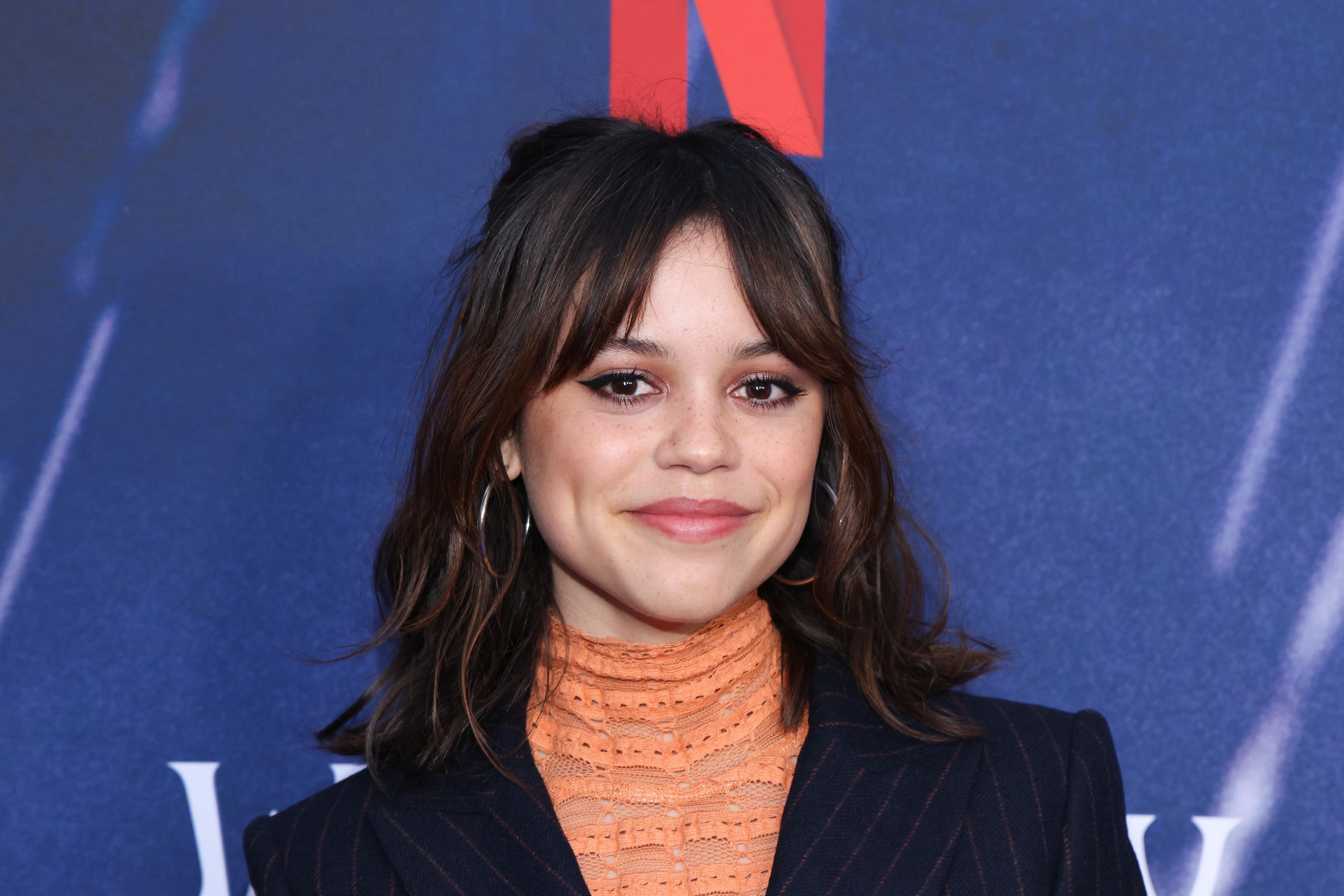 Jenna Ortega smiles in front of a Netflix backdrop, wearing a high-neck lace top and a pinstriped blazer