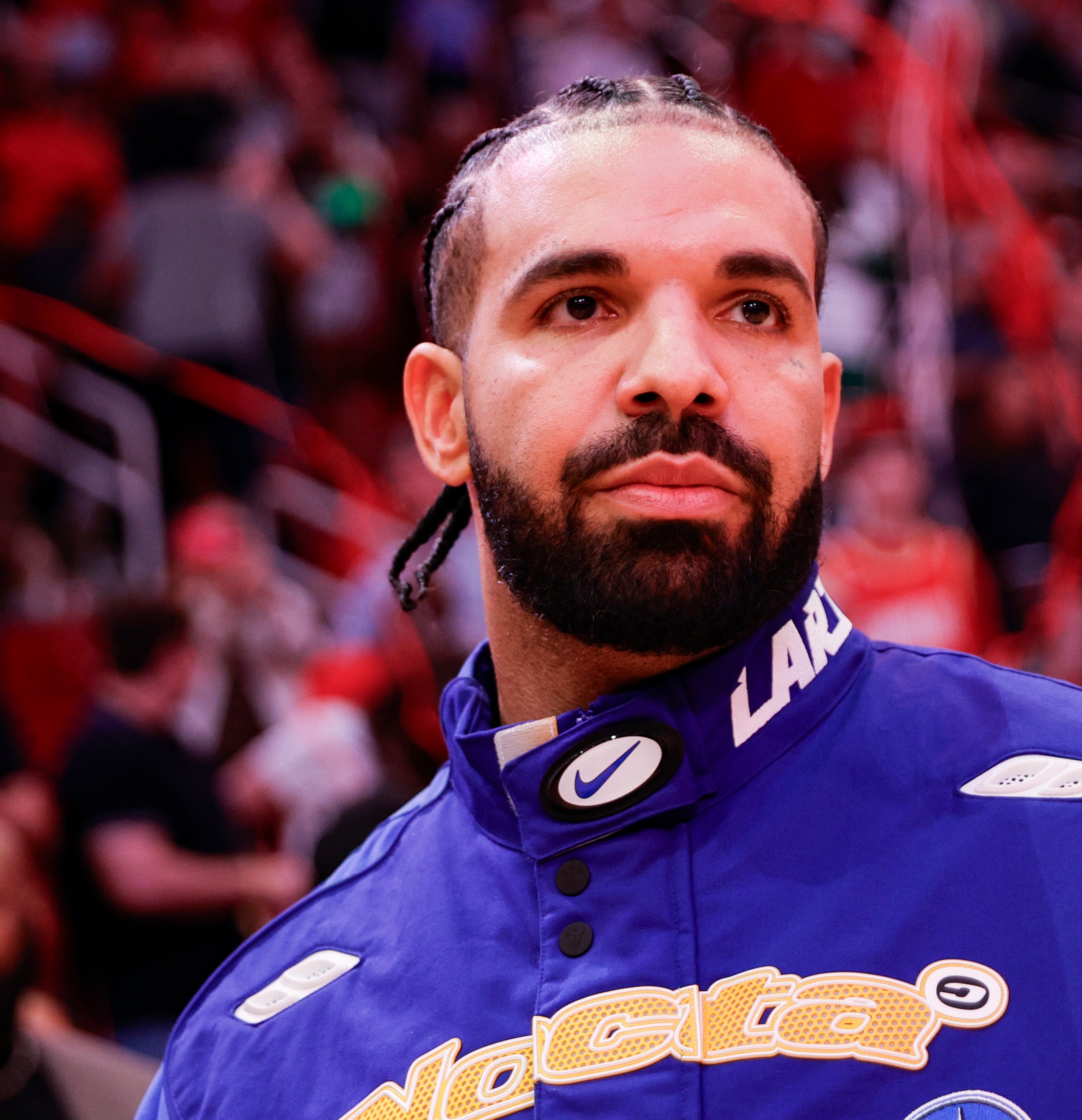 Drake stands in a sports venue wearing a blue Nocta jacket with a focused expression, looking ahead