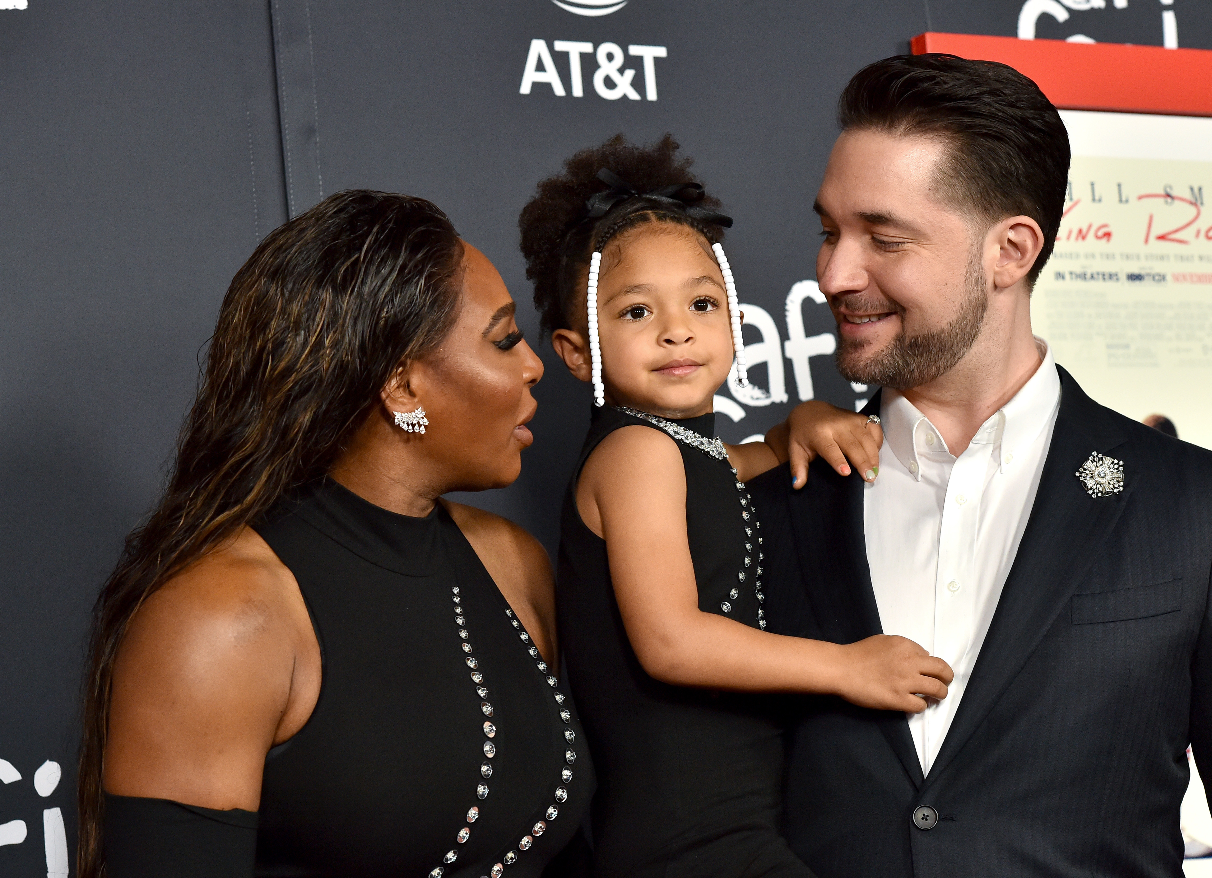 Serena Williams, Alexis Olympia Ohanian Jr., and Alexis Ohanian at a red carpet event. Serena and Alexis Jr. wear stylish black outfits, while Alexis wears a suit