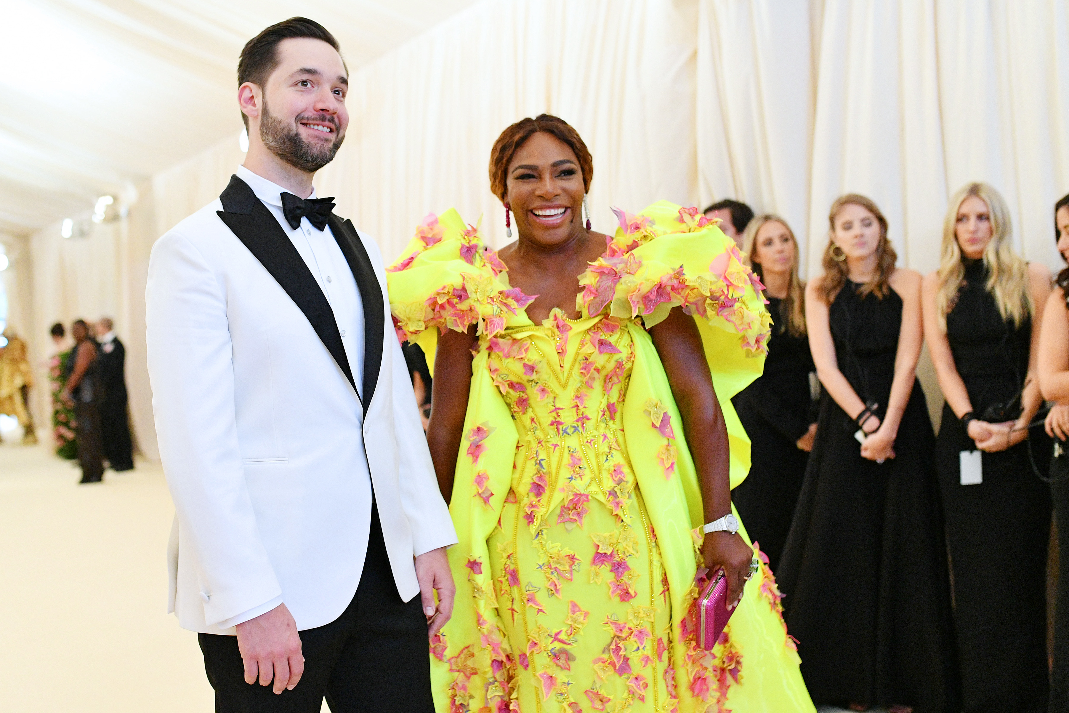 Alexis Ohanian in a white tuxedo with black bowtie and Serena Williams in an extravagant yellow and pink floral dress at an event, with six onlookers in black attire in the background