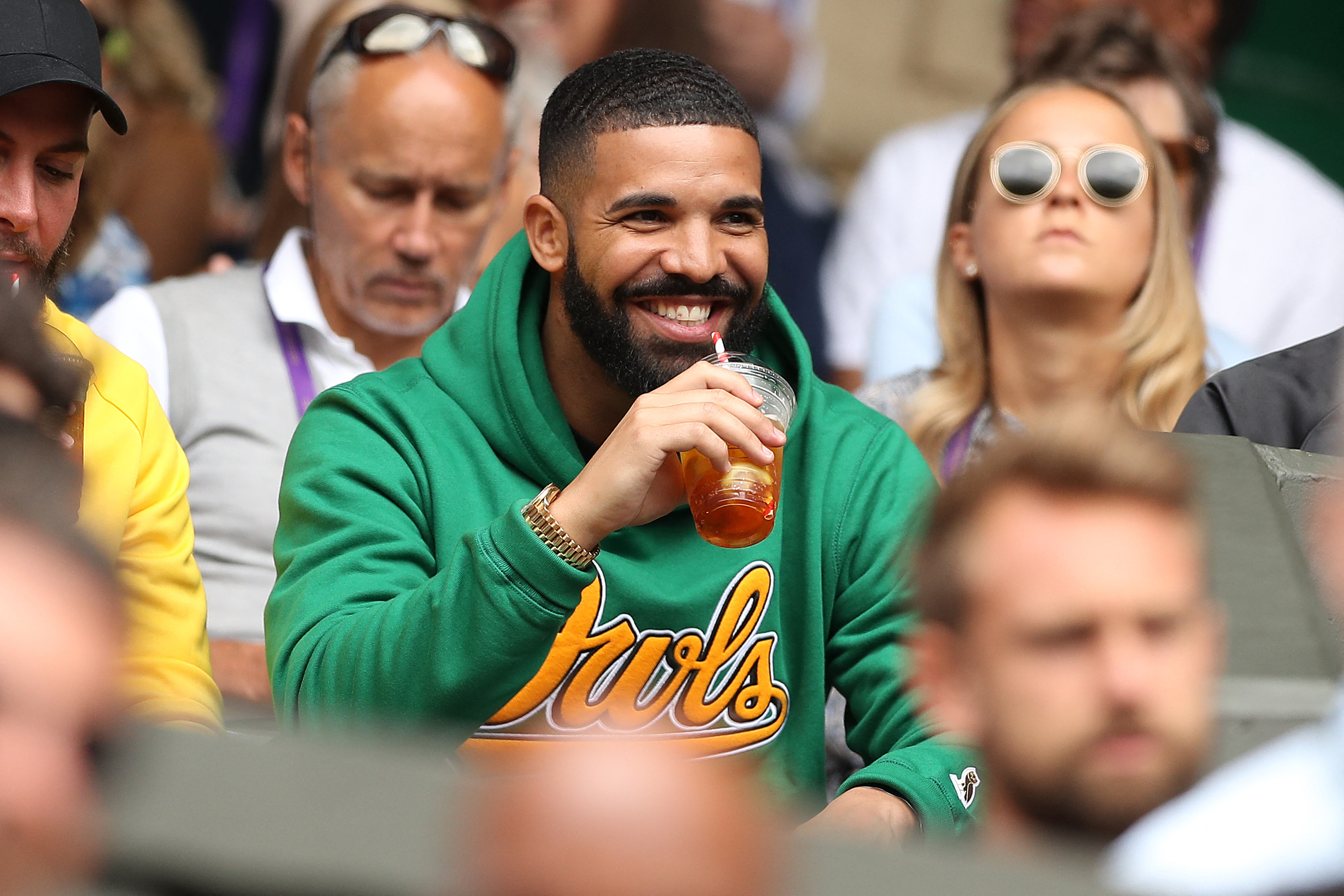 Celebrity Drake enjoys a drink while seated in a crowd at an event, wearing a green hoodie