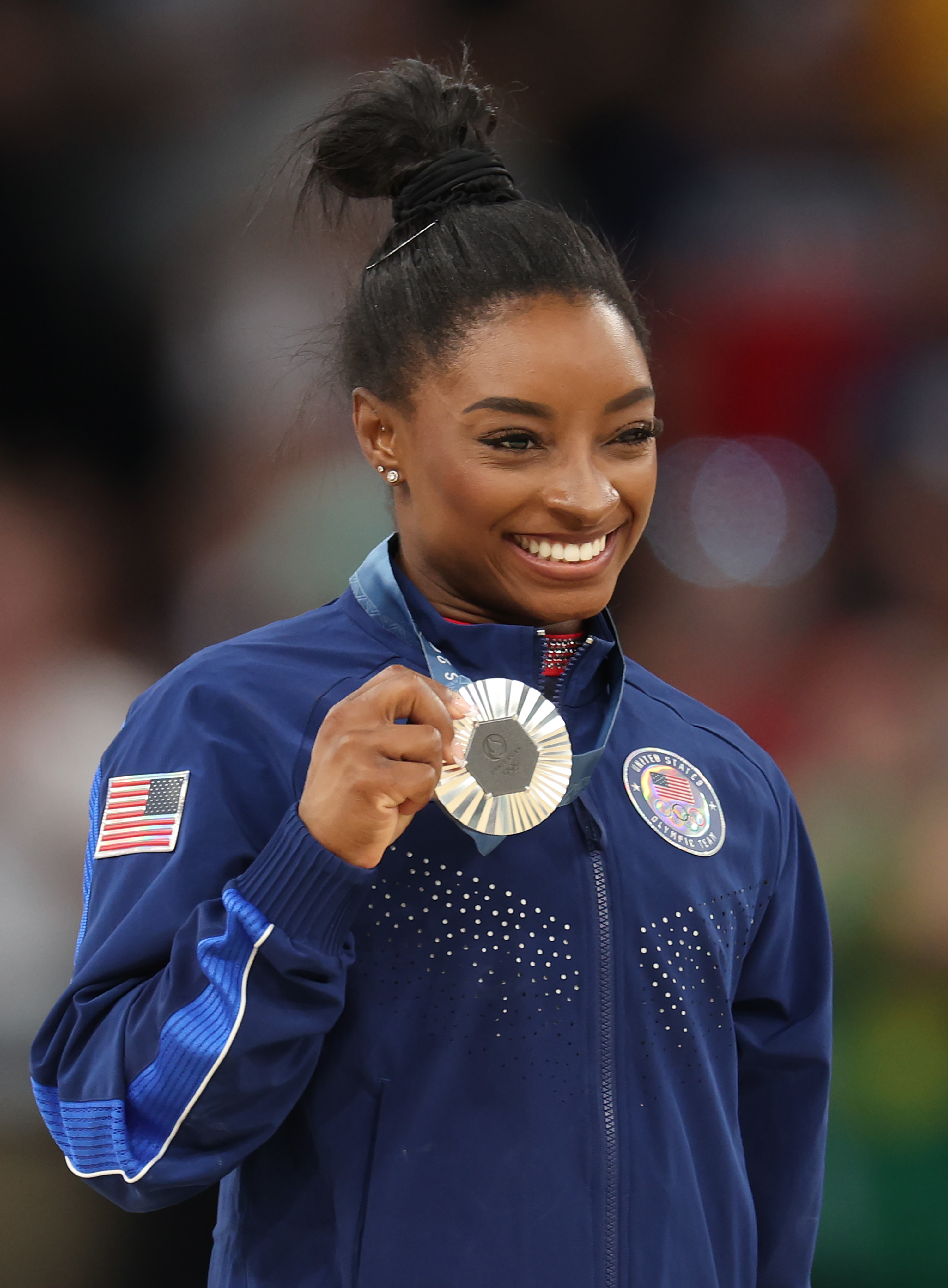 Simone Biles smiles proudly while holding her silver medal at a gymnastic event, wearing a team USA tracksuit with the American flag patch on the sleeve