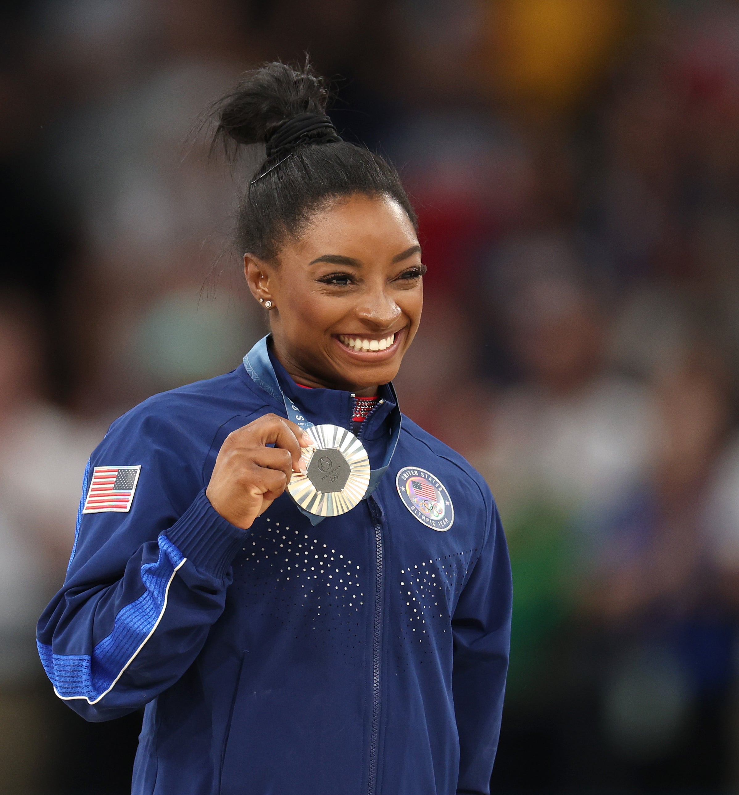 Simone Biles smiles and holds up her silver medal while wearing a USA team tracksuit at an event