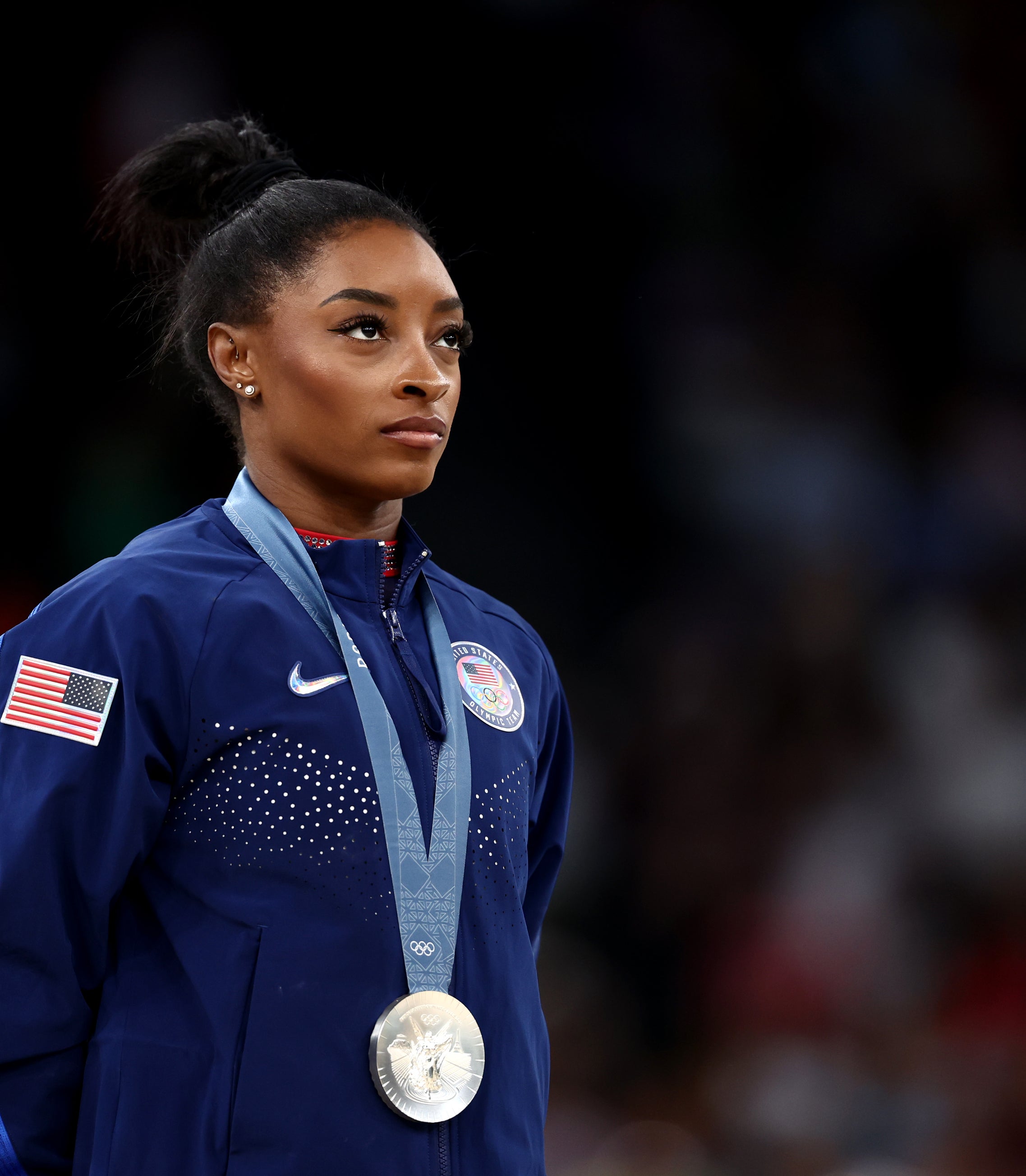 Simone Biles stands, wearing a Team USA jacket with a silver medal around her neck. She looks focused and determined