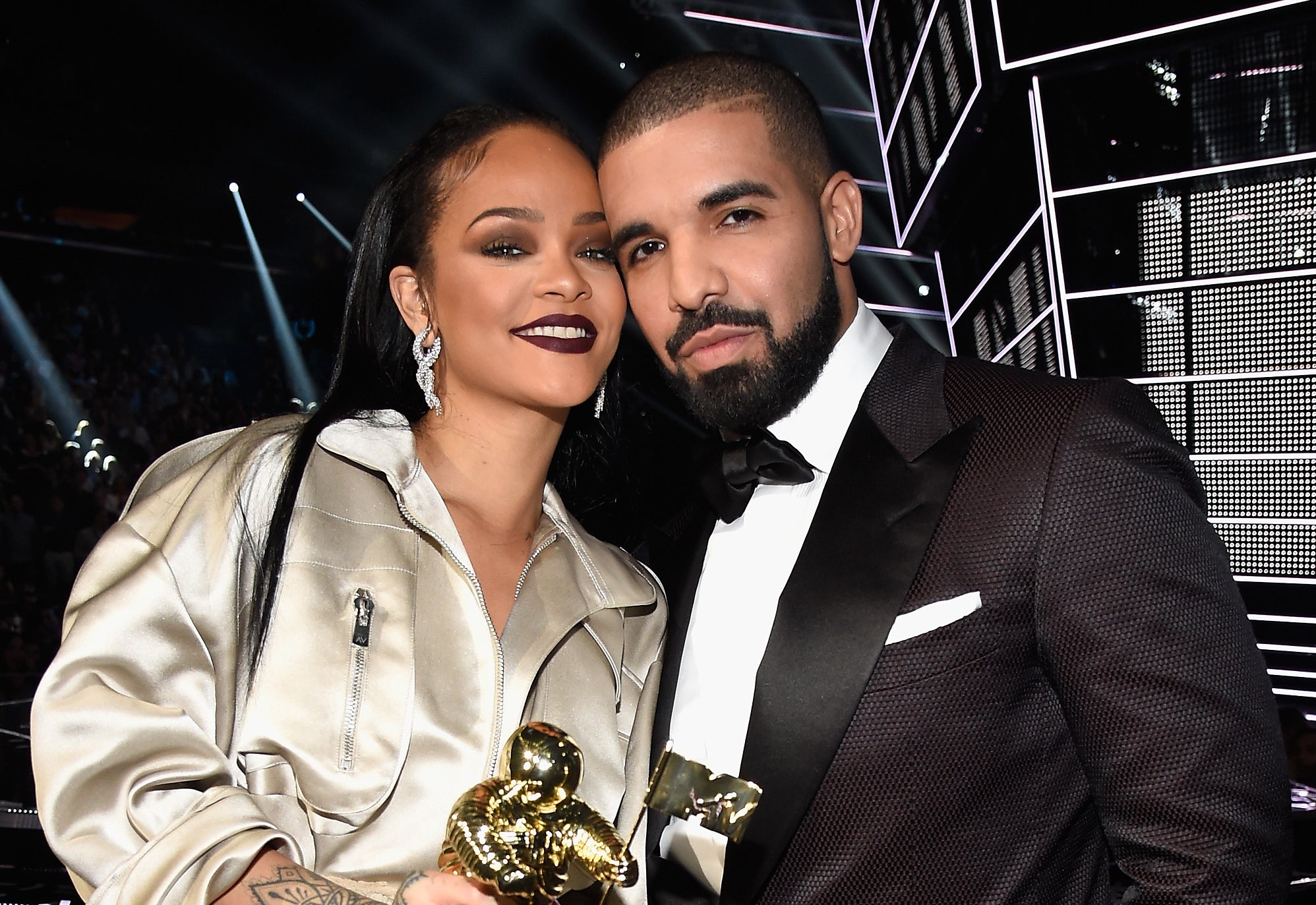 Rihanna holds an award and stands next to Drake. Rihanna wears a long coat, and Drake is in a tuxedo. They are on stage at an event