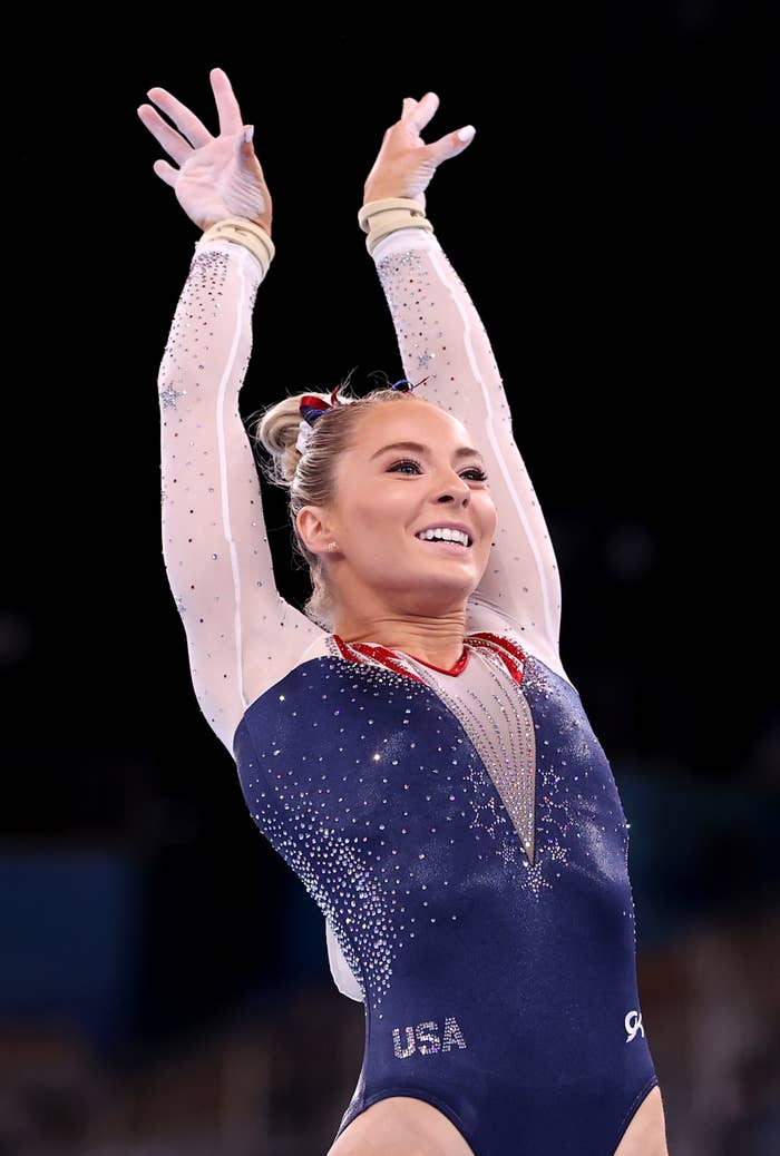 A gymnast is performing a routine with arms raised, smiling confidently. She is wearing a sparkling leotard with "USA" written on it