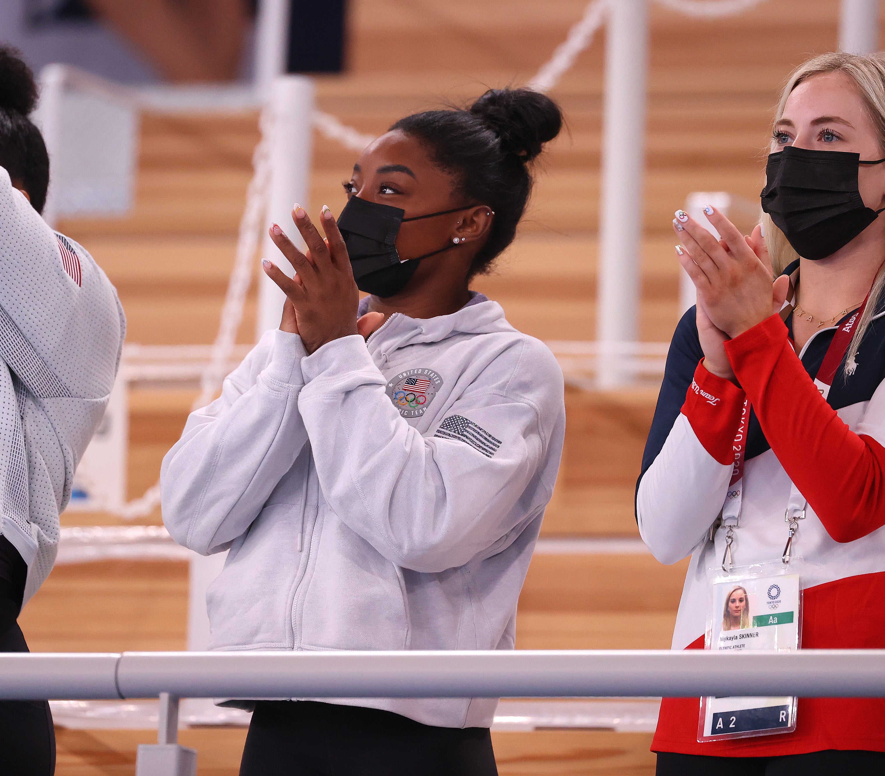 Simone Biles and MyKayla Skinner clap and cheer while wearing masks. They are standing in an indoor venue, dressed in team sportswear