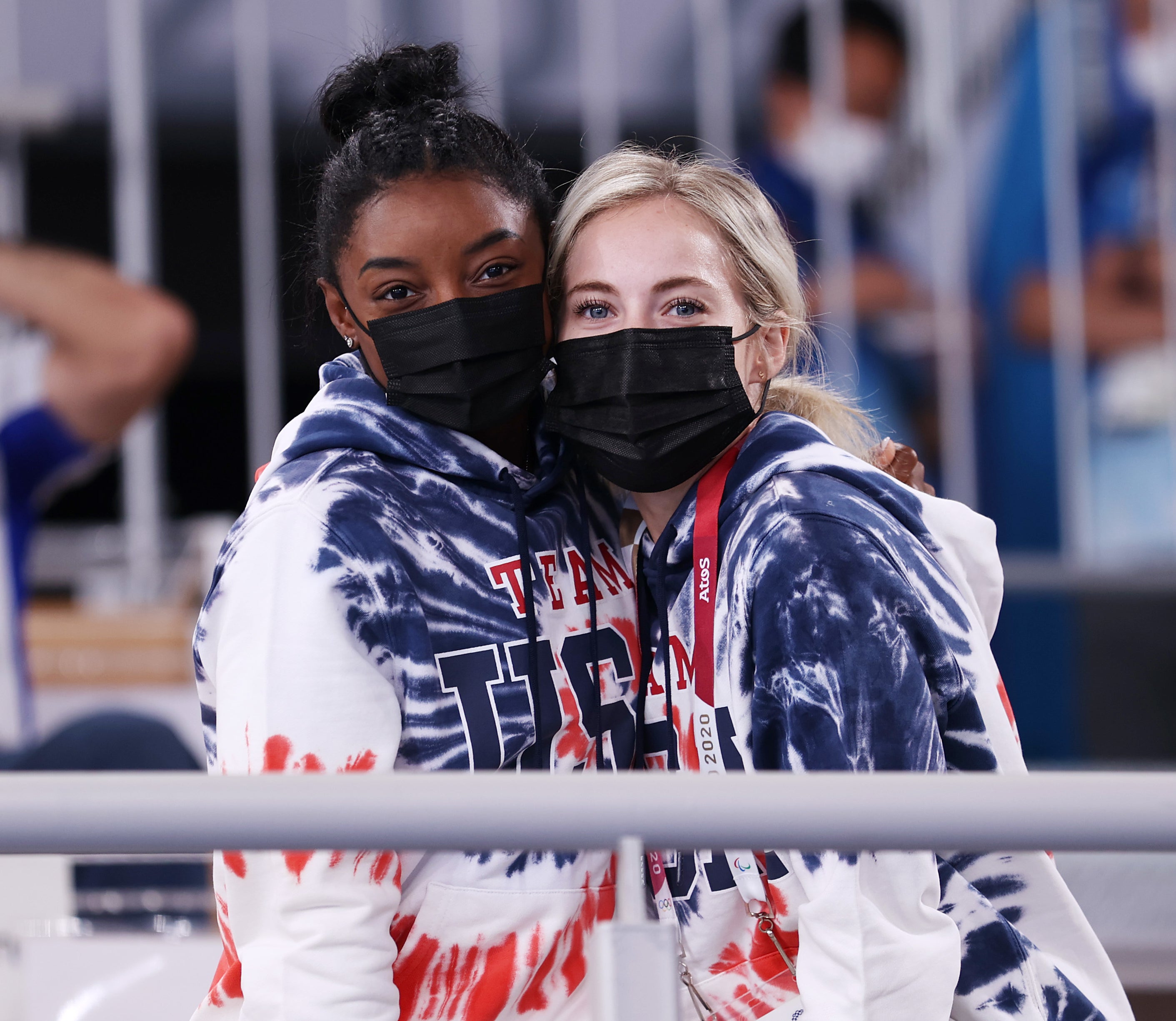 Simone Biles and MyKayla Skinner, wearing Team USA tie-dye hoodies and black masks, hug each other while standing in a stadium