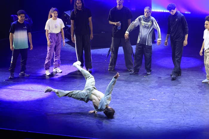 A group of eight dancers watches a breakdancer spinning on his shoulders onstage. The scene is lit by stage lights