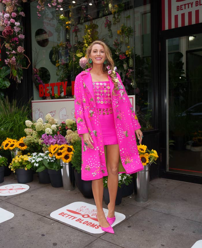 Blake Lively stands outside Betty Blooms in a stylish floral-adorned outfit with matching pink coat, skirt, top, and heels, surrounded by flowers and plants