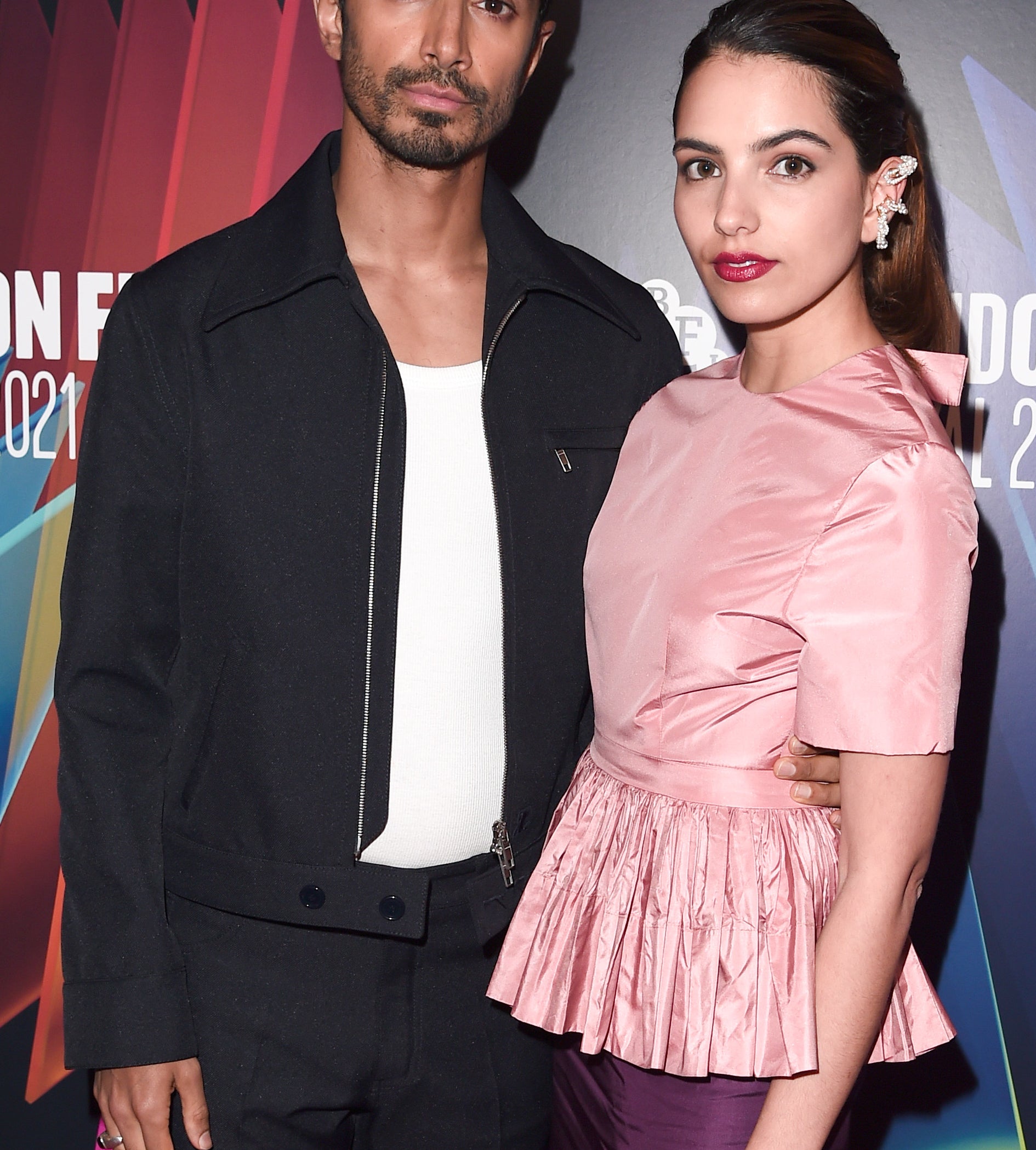 Riz Ahmed and Fatima Farheen Mirza pose at the London Film Festival 2021. Riz wears a black jacket over a white shirt, and Fatima wears an elegant pink silk dress