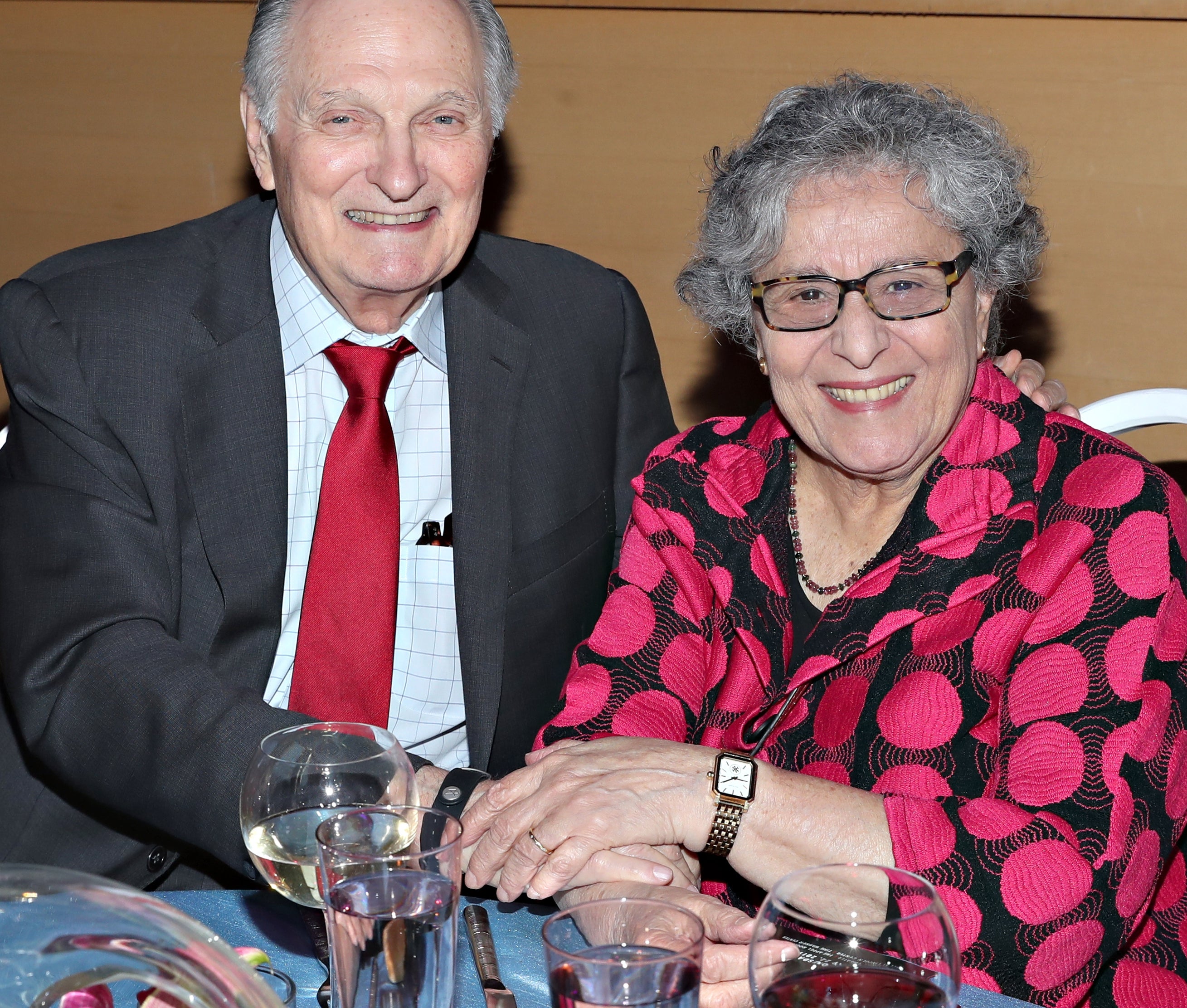 Alan Alda in a suit and tie, sits beside Arlene Alda in a patterned blouse at a table with drinks and decorations, both smiling at the camera